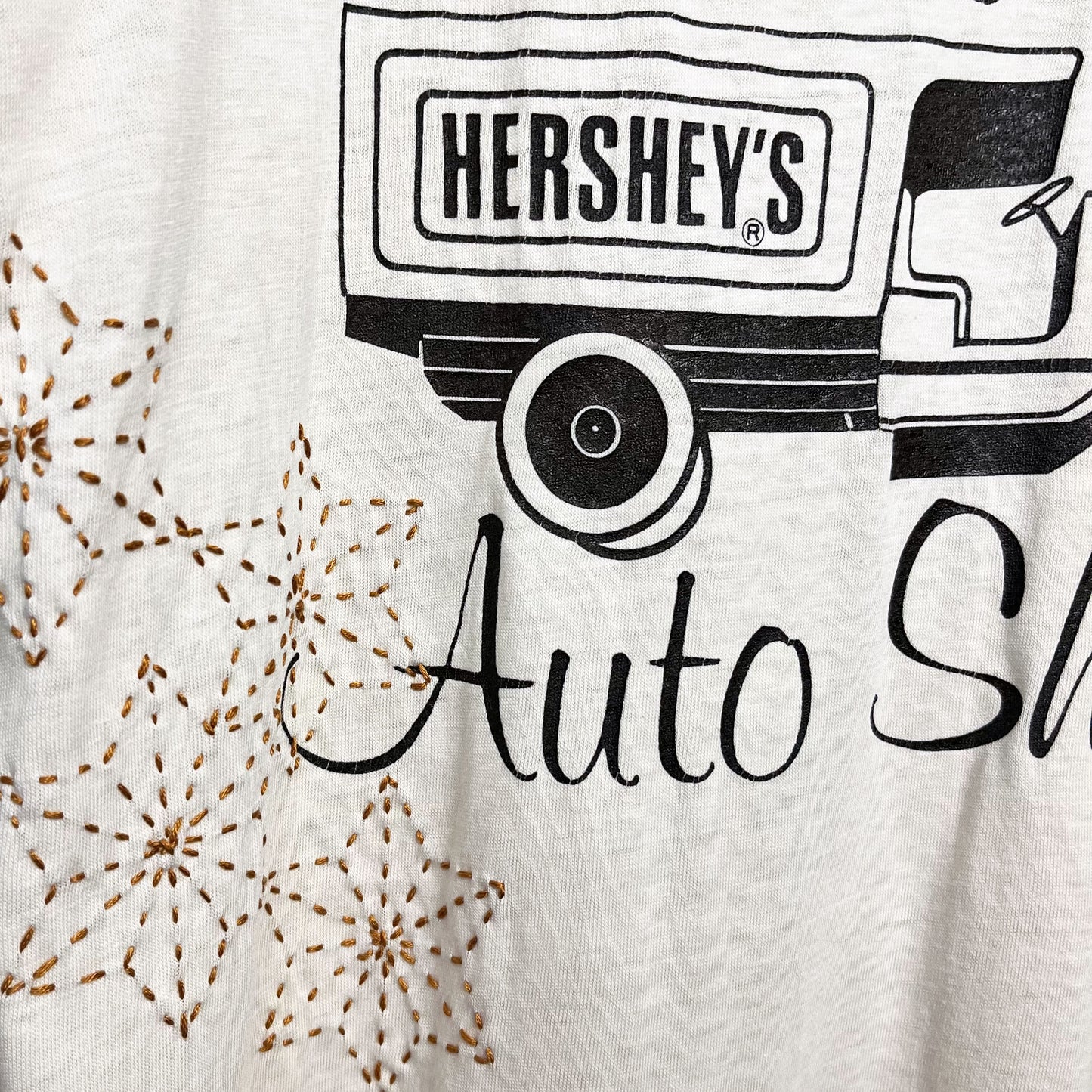 Close up view of a cream colored T-shirt, the t-shirt has a graphic print of a truck that says Hershey's on the back, and the word Auto below the truck, next to the word Auto there are hand embroidered sashiko stars in mustard brown
