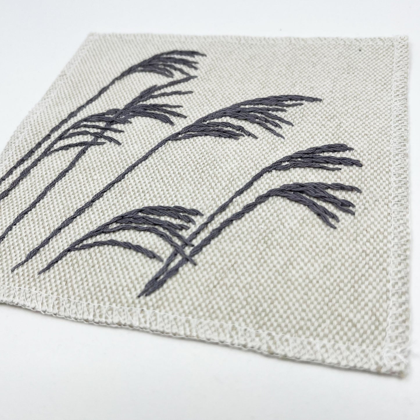 close up angled view of a cream colored square patch with stalks of wild grass stitched in grey thread with a stem stitch, on a white background