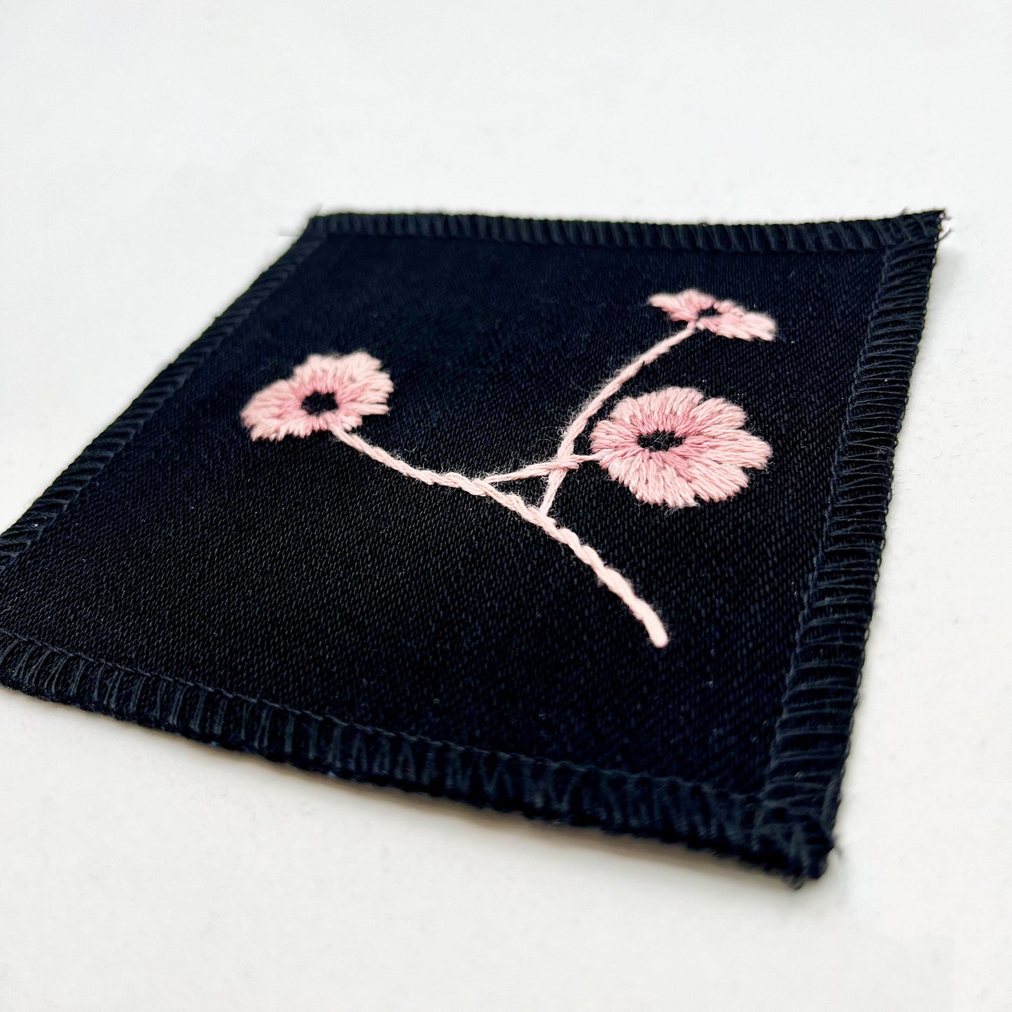 a close up angled view of a black colored square patch, hand embroidered with three poppies on a stem, in shades of pink, with overlocked edges, on a white background