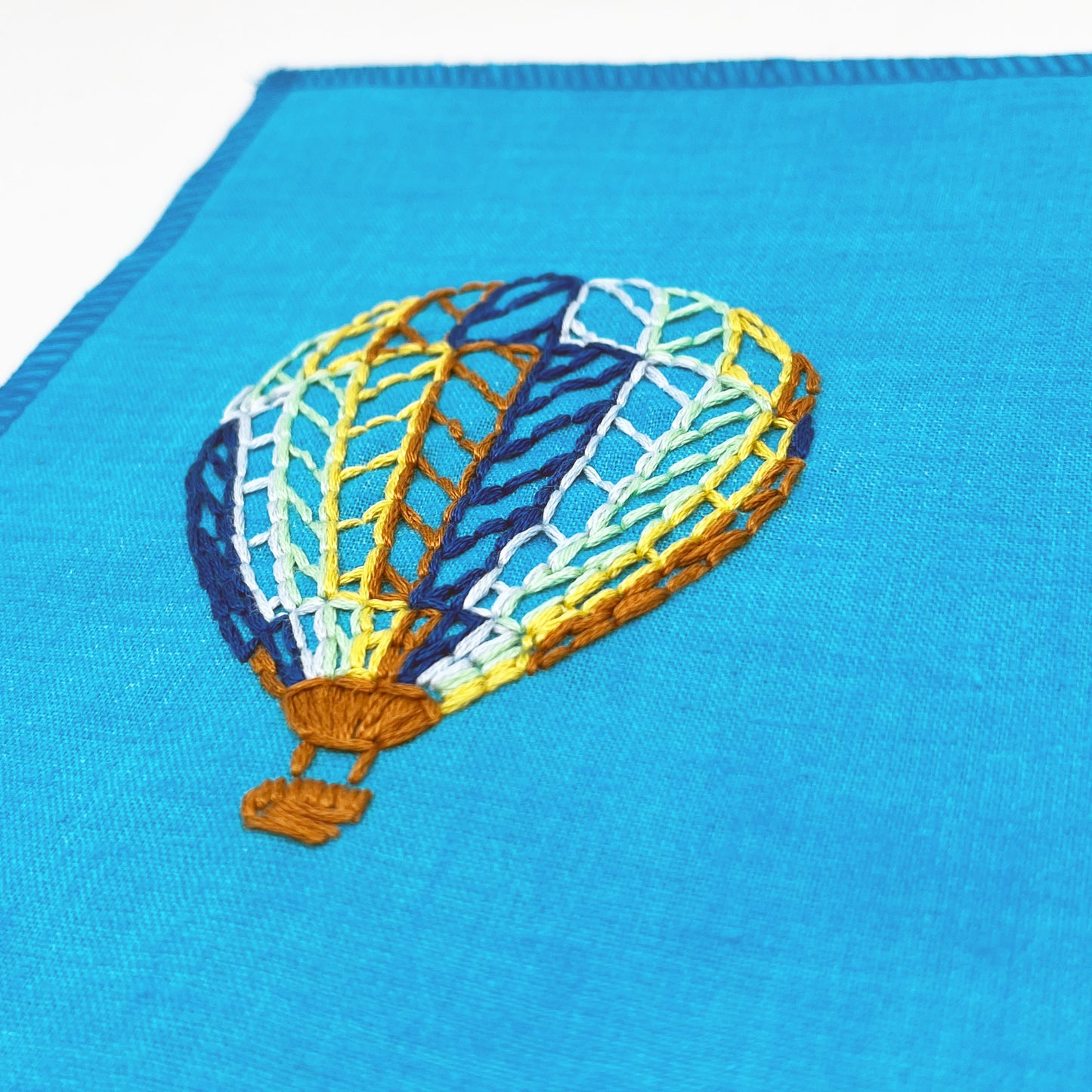 a close up angled view of a bright blue fabric wall hanging with a hot air balloon stitched on it off center, in dark blue, light blue, light green, yellow and rust colored thread, on a white background