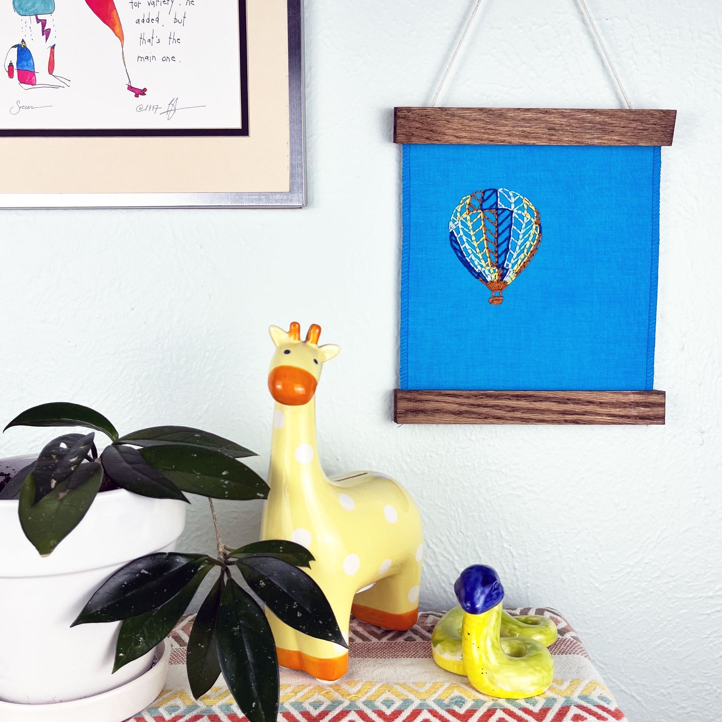 a bright blue fabric wall hanging in a magnetic wood frame, with a hot air balloon stitched on it off center, in dark blue, light blue, light green, yellow and rust colored thread, hanging on a wall near other colorful artwork and a giraffe coin bank