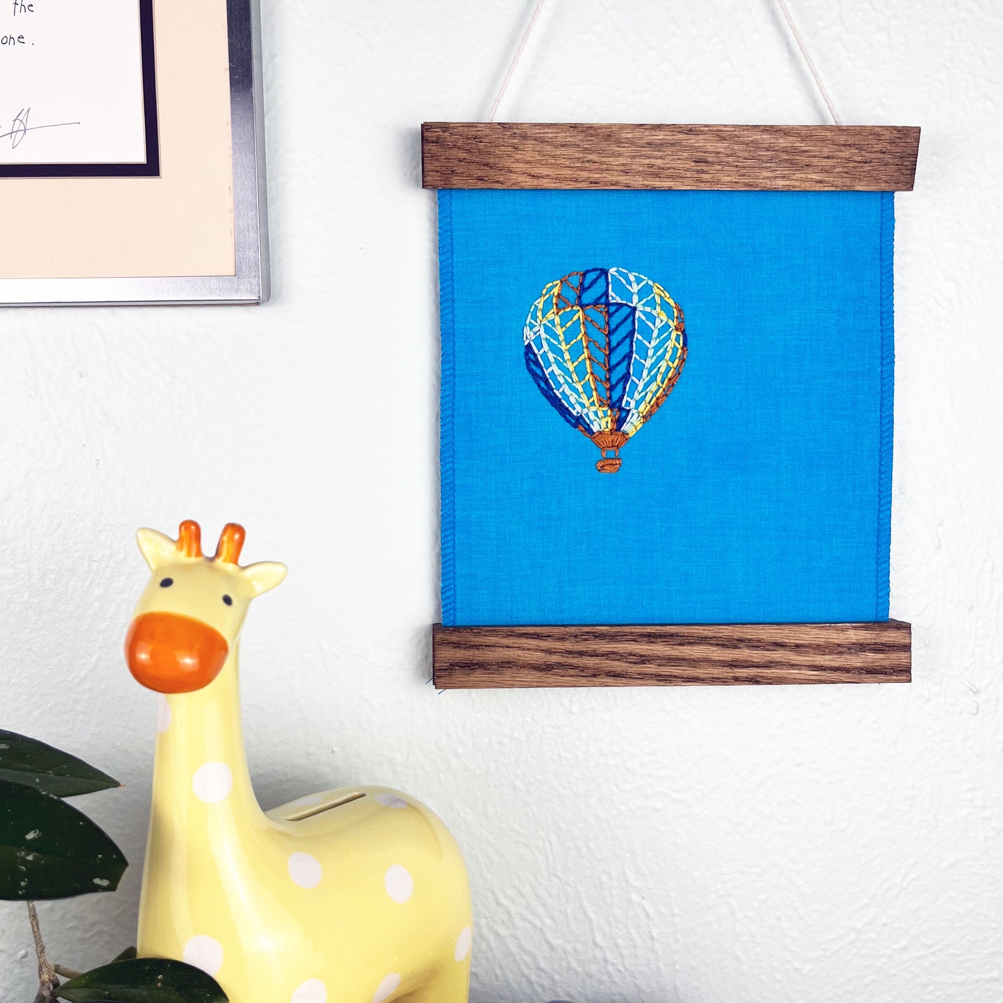 a bright blue fabric wall hanging in a magnetic wood frame, with a hot air balloon stitched on it off center, in dark blue, light blue, light green, yellow and rust colored thread, hanging on a wall near other colorful artwork and a giraffe coin bank