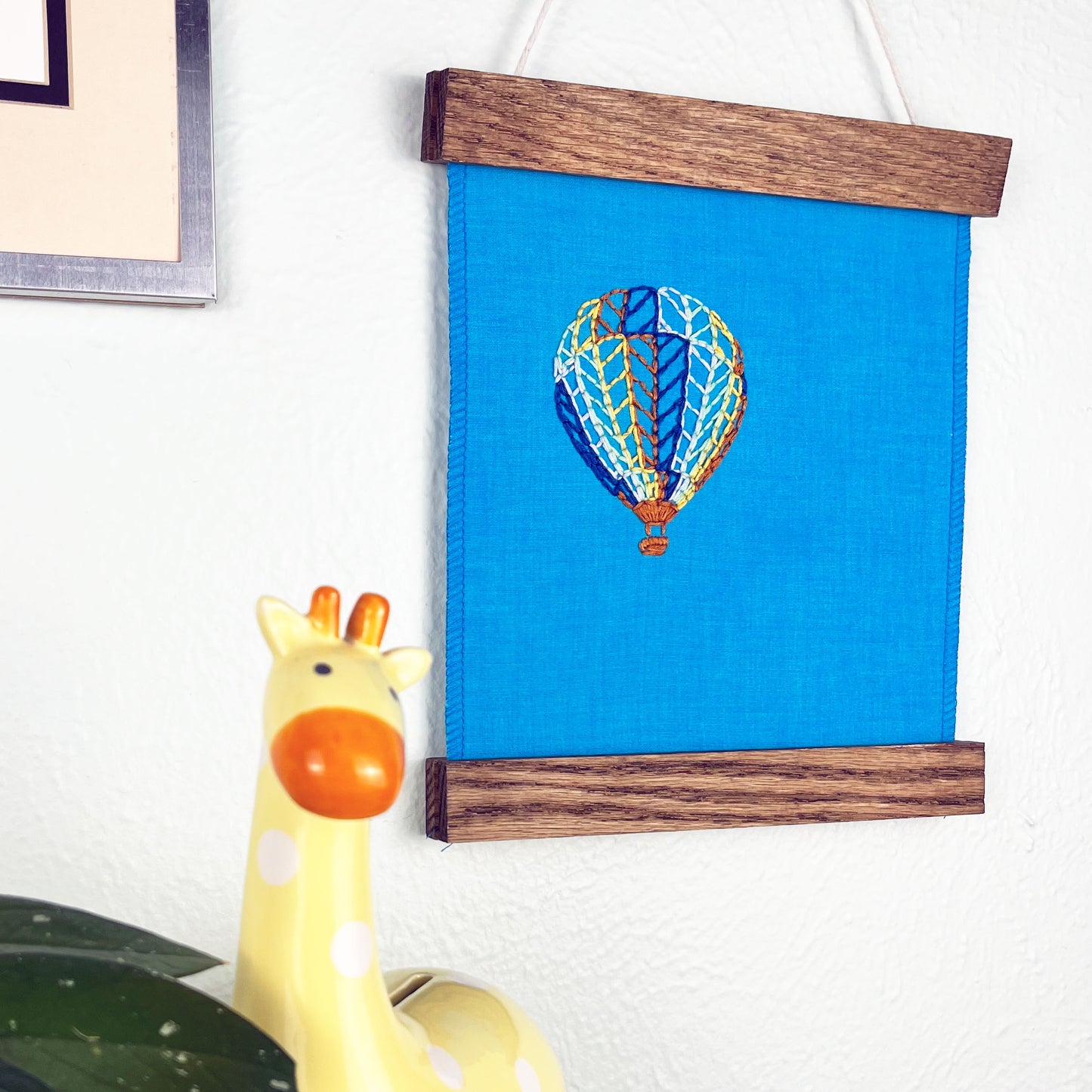 a bright blue fabric wall hanging in a magnetic wood frame, with a hot air balloon stitched on it off center, in dark blue, light blue, light green, yellow and rust colored thread, hanging on a wall near other colorful artwork and a giraffe coin bank