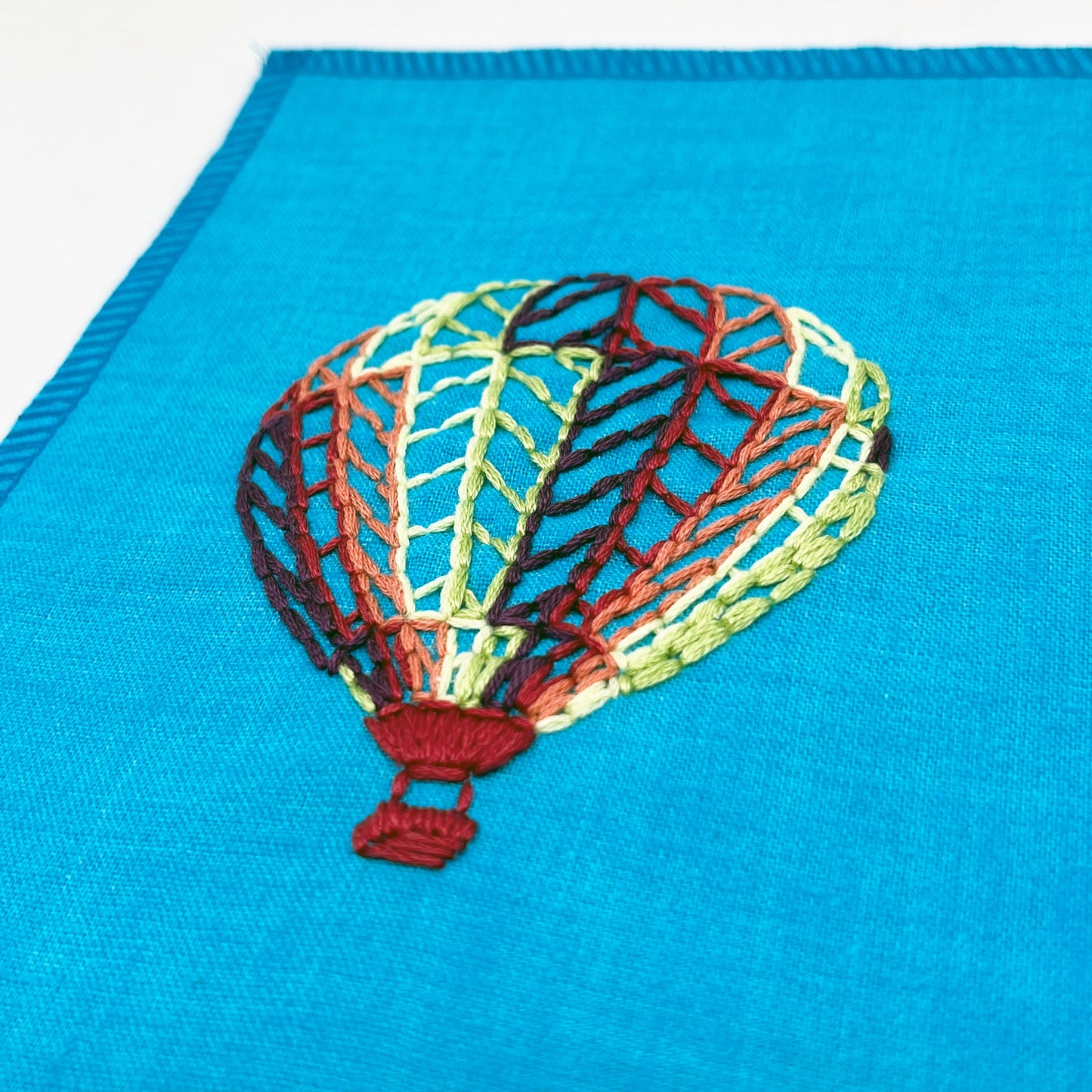 close up angled view of a bright blue fabric wall hanging with a hot air balloon stitched on it off center, in plum, magenta, coral, lime green and yellow colored thread