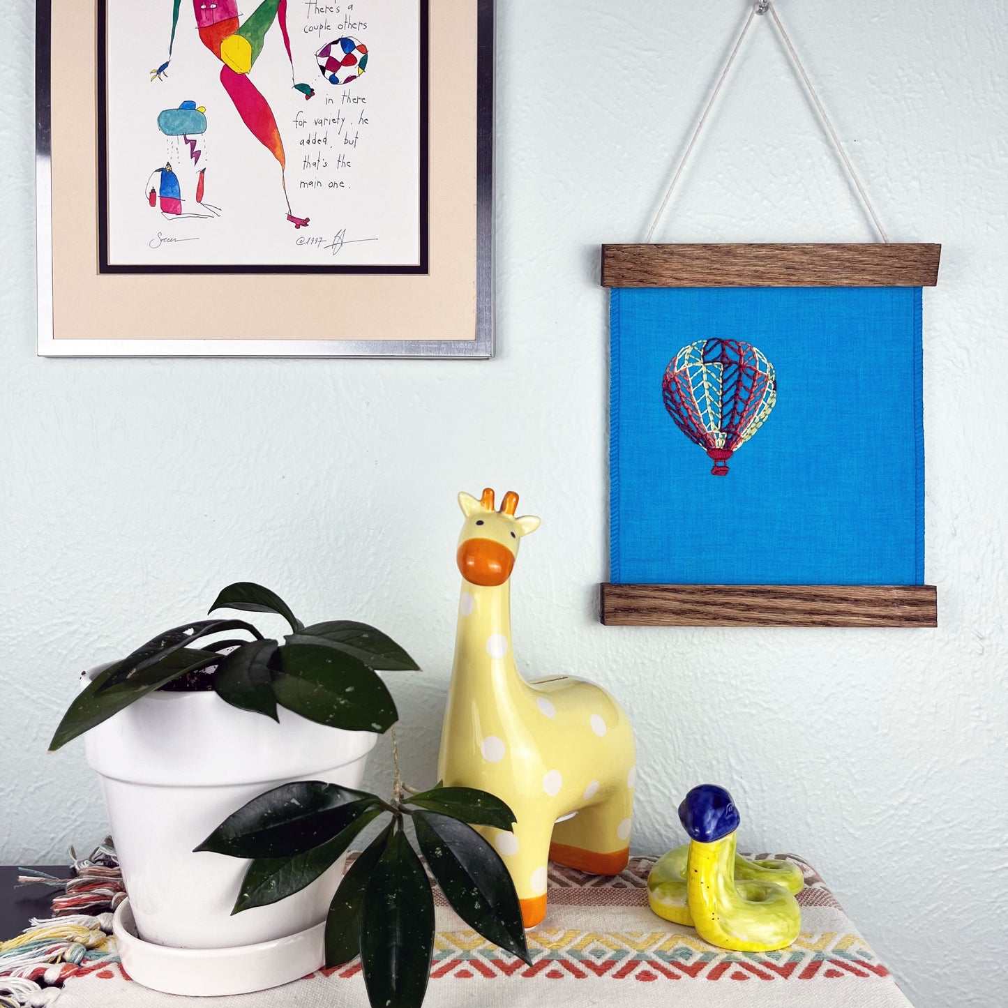 a bright blue fabric wall hanging in a magnetic wood frame, with a hot air balloon stitched on it off center, in plum, magenta, coral, lime green and yellow colored thread, hanging on a wall near other colorful artwork