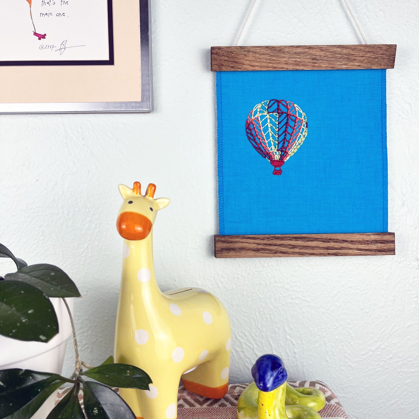 a bright blue fabric wall hanging in a magnetic wood frame, with a hot air balloon stitched on it off center, in plum, magenta, coral, lime green and yellow colored thread, hanging on a wall near other colorful artwork