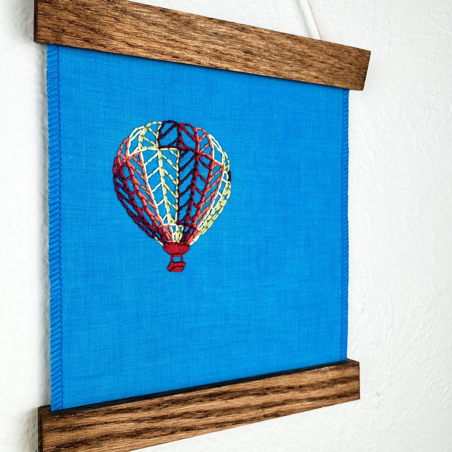 close up angled view of a bright blue fabric wall hanging in a magnetic wood frame, with a hot air balloon stitched on it off center, in plum, magenta, coral, lime green and yellow colored thread, hanging on a wall