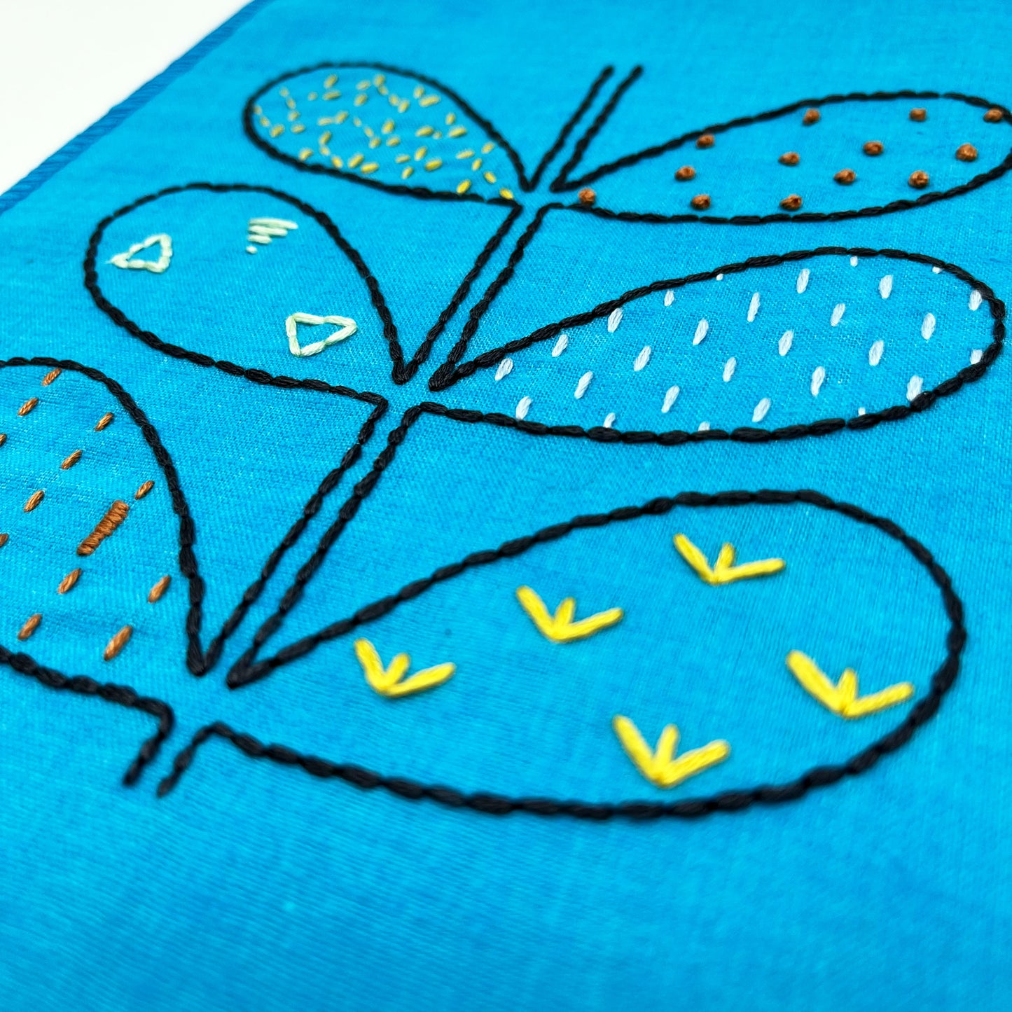 close up view of a small bright blue fabric wall hangings, hand embroidered with the outline of symmetrical petals on a stem in navy, each petal filled with different stitches like french knots, running stitches and triangles, in different colors