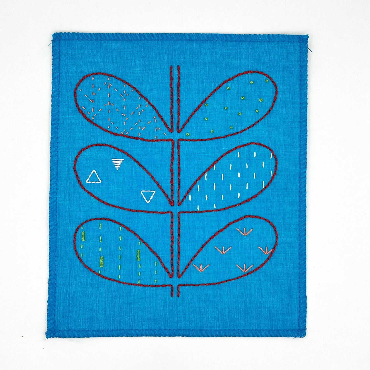 small bright blue fabric wall hangings, hand embroidered with the outline of symmetrical petals on a stem in red, each petal filled with different stitches like french knots, running stitches and triangles, in different colors, on a white background