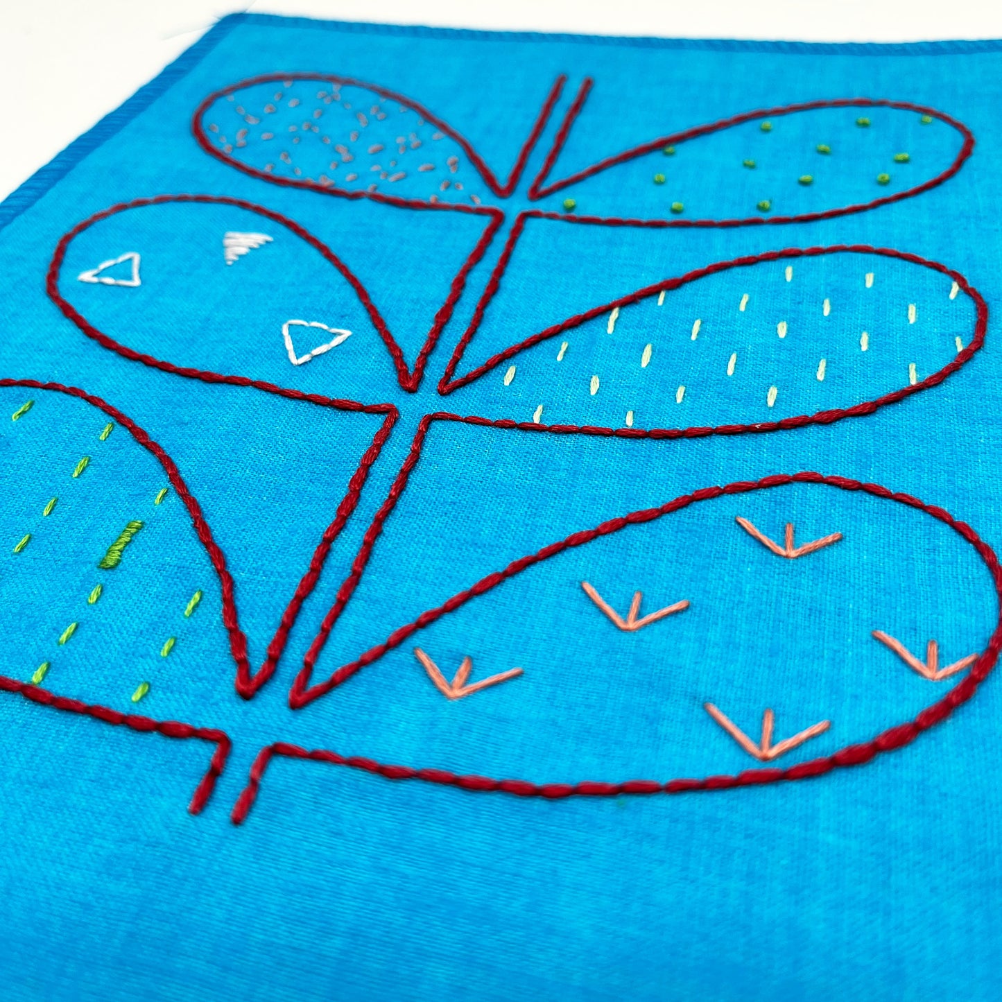 close up view of a small bright blue fabric wall hangings, hand embroidered with the outline of symmetrical petals on a stem in red, each petal filled with different stitches like french knots, running stitches and triangles, in different colors