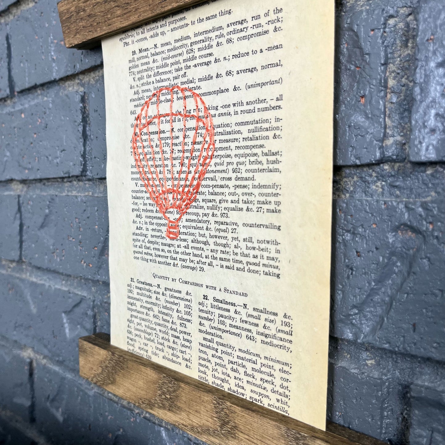 a close up angled view of a vintage thesaurus page, hand stitched over with an outline of a hot air balloon in coral thread, hanging in a magnetic wood frame on a grey brick wall