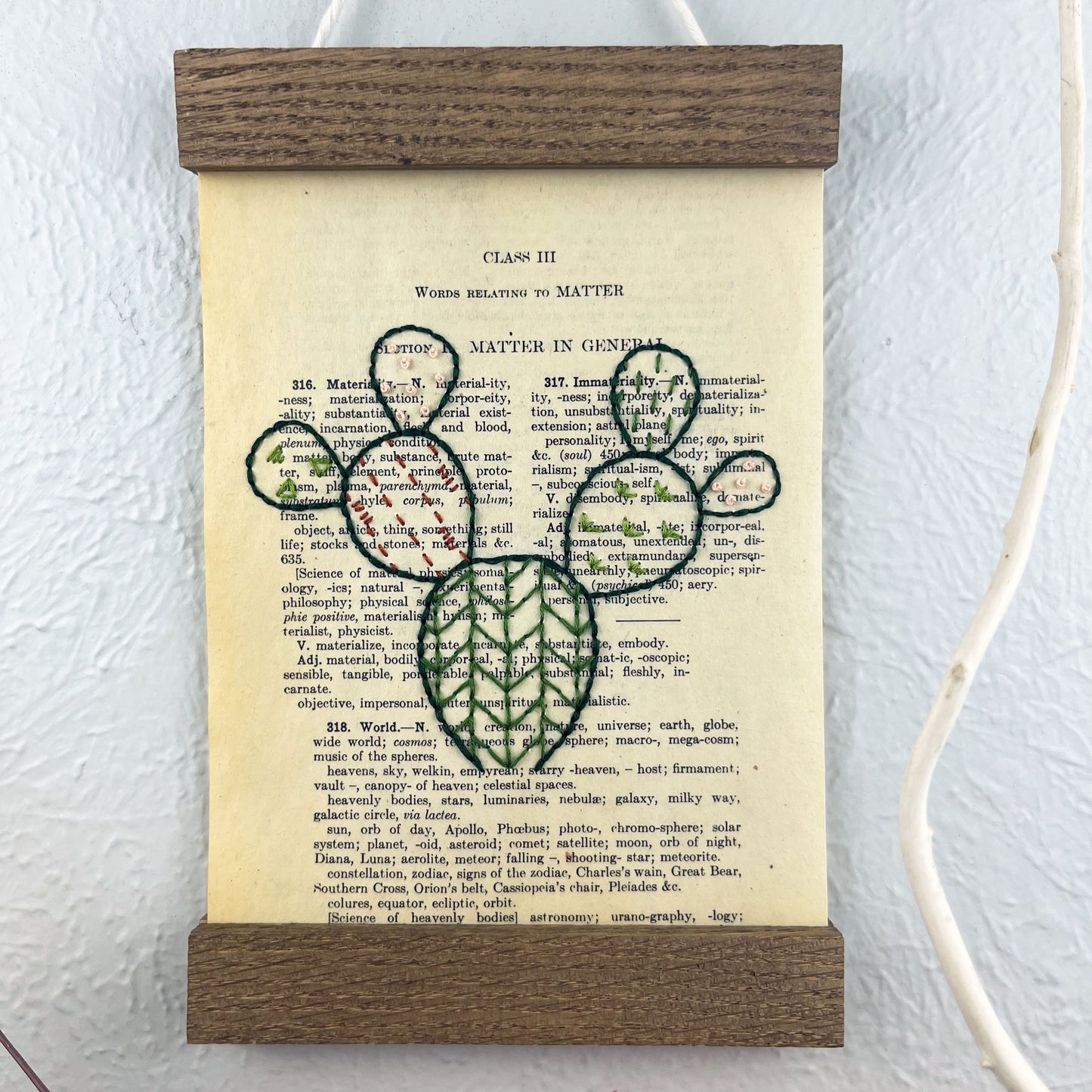 close up view of a vintage thesaurus page in a magnetic wood frame hanging on a wall, the book page is hand embroidered in green peach and rust colored thread with a prickly pear cactus with different patterns filled in each pad