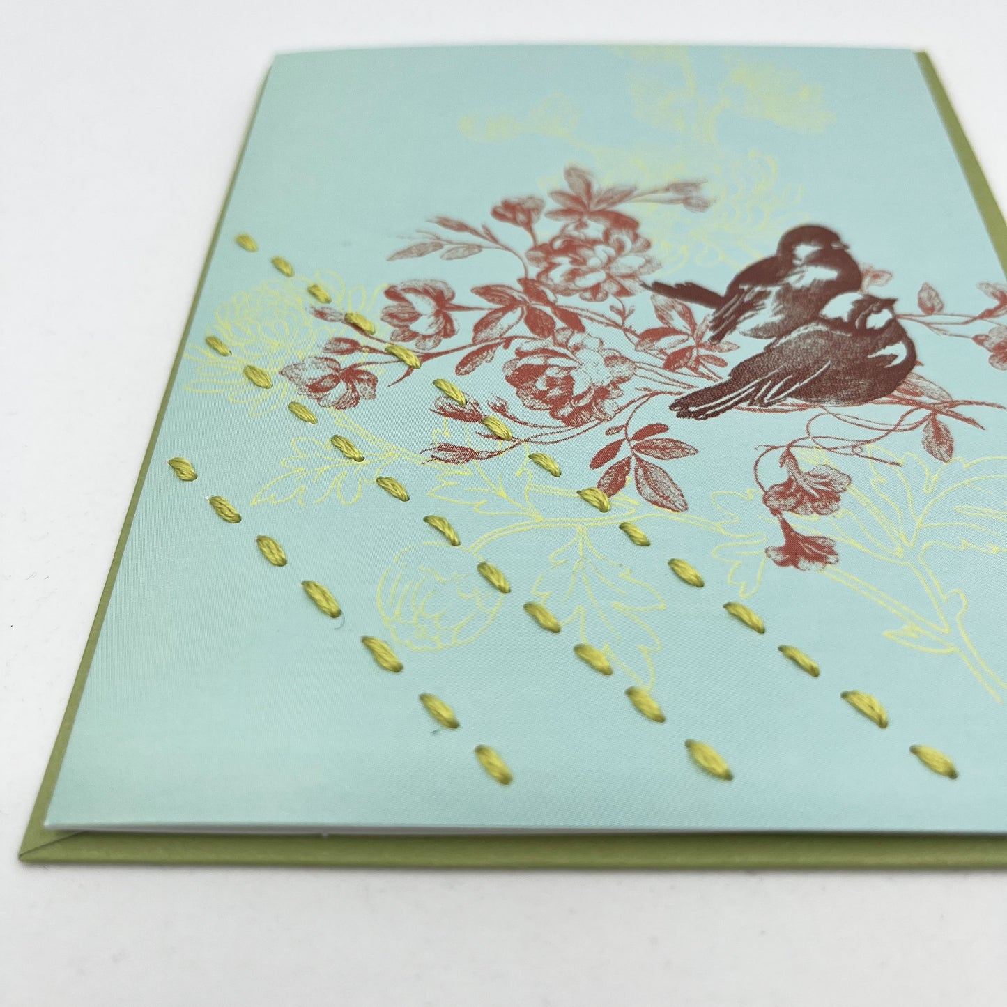 a close up angled view of a robin's egg blue greeting card on a green envelope with a sparrow and flowers printed on it in reds and chartreuse, hand stitched over with 3 rows of running stitches in chartreuse, diagonally over the bottom left corner