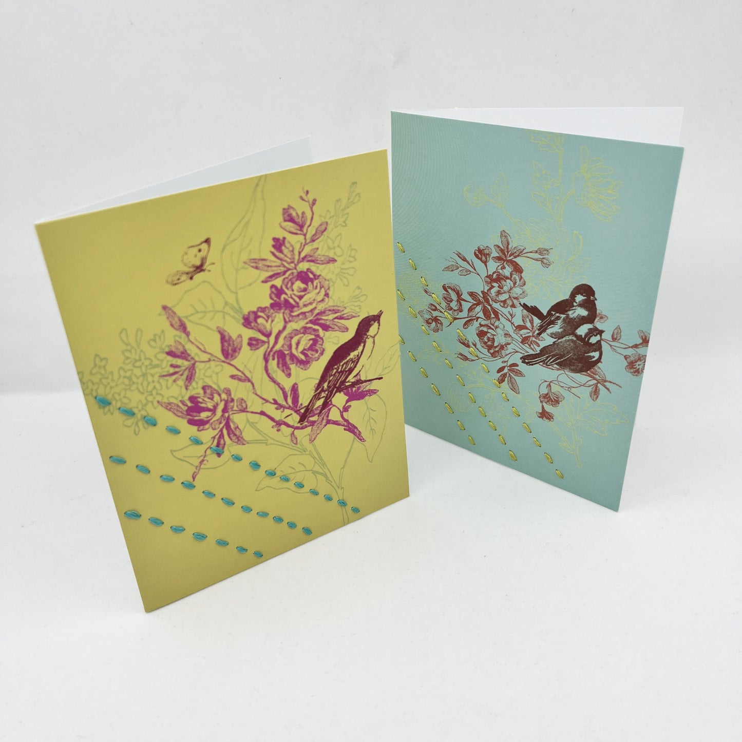 two greeting cards, one chartreuse and one robin's egg blue, standing upright, with a sparrow and flowers printed on it in brick red and fuchsia, hand stitched over with 3 rows of running stitches, diagonally over the bottom left corner