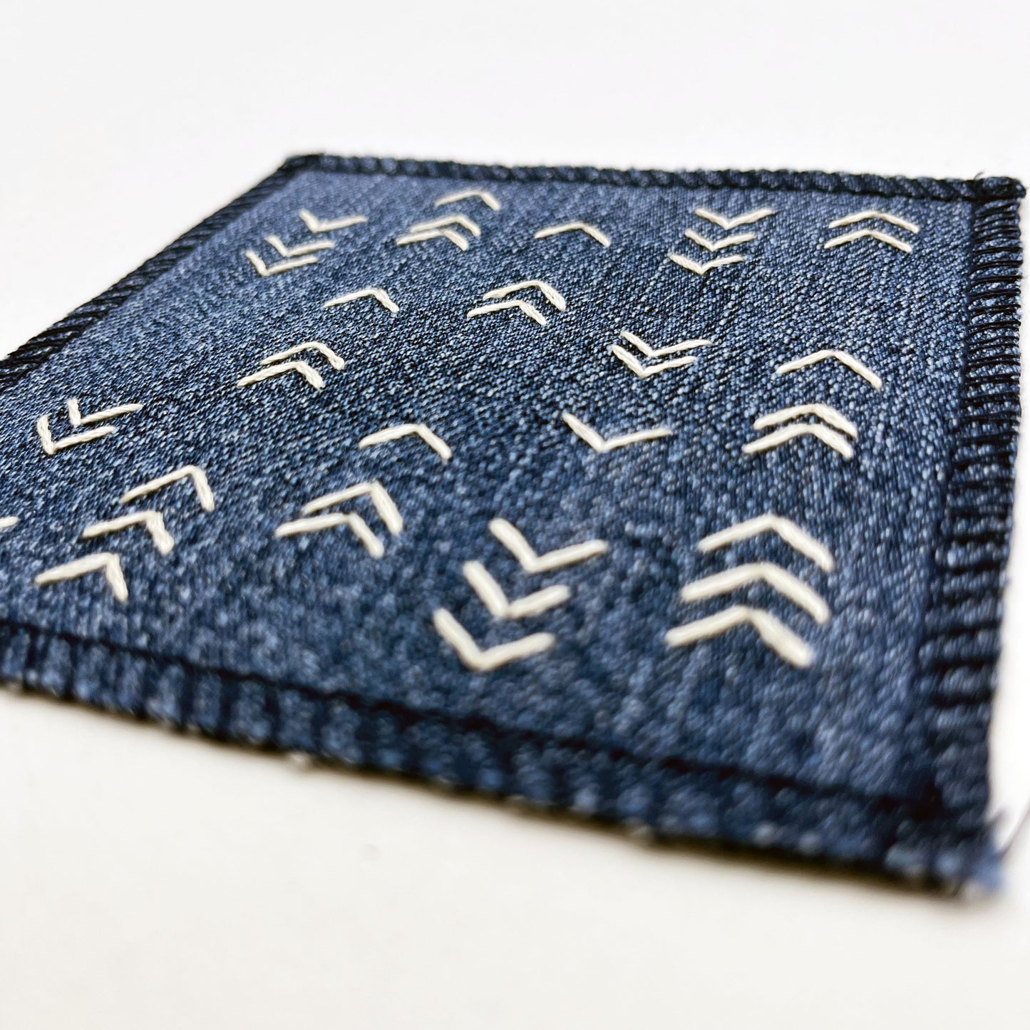 close up angled view of a square denim patch hand stitched in ivory with rows of unevenly spaced small chevrons facing different directions on a white background
