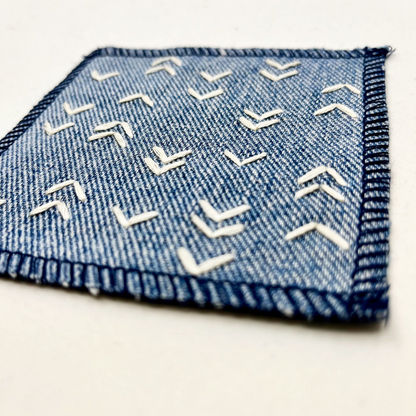 close up angled view of a square denim patch hand stitched in ivory with rows of unevenly spaced small chevrons facing different directions on a white background