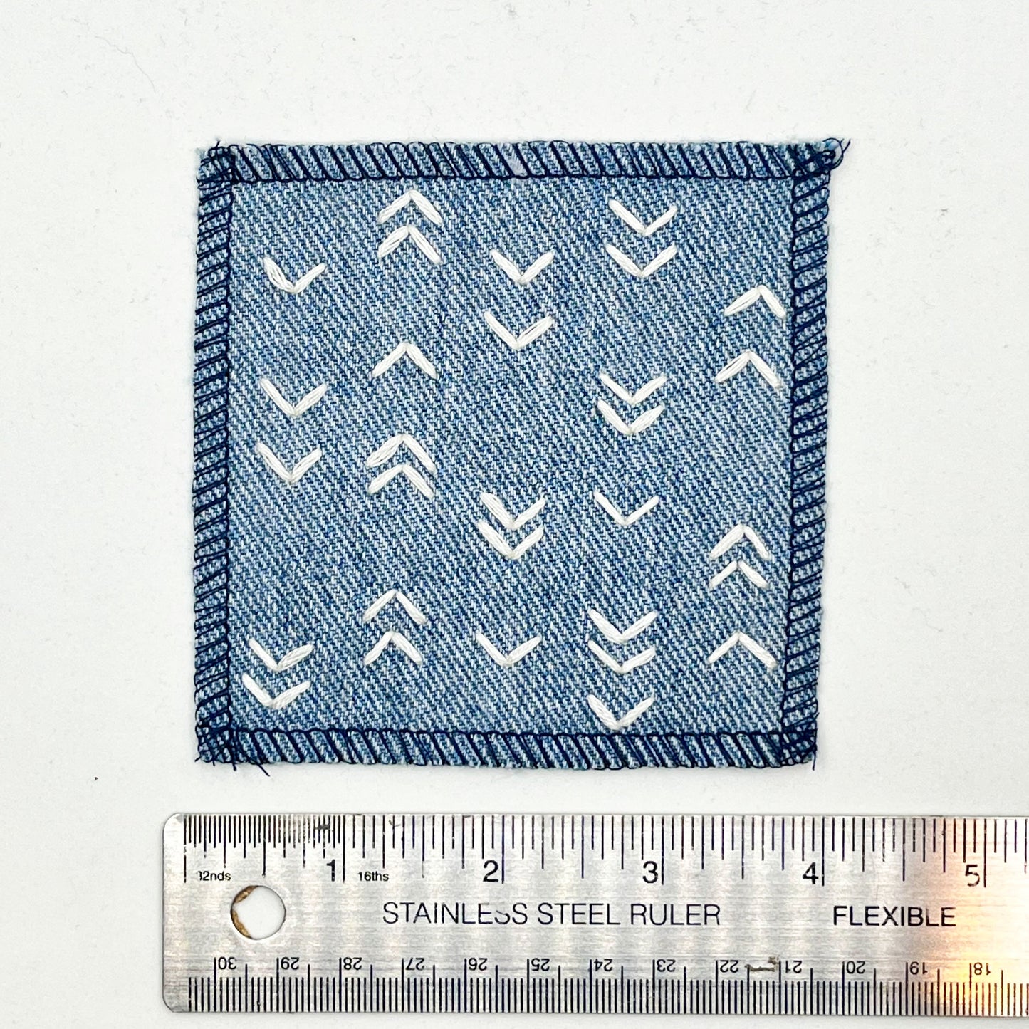 a square denim patch hand stitched in ivory with rows of unevenly spaced small chevrons facing different directions placed next to a metal ruler to show a width of four inches