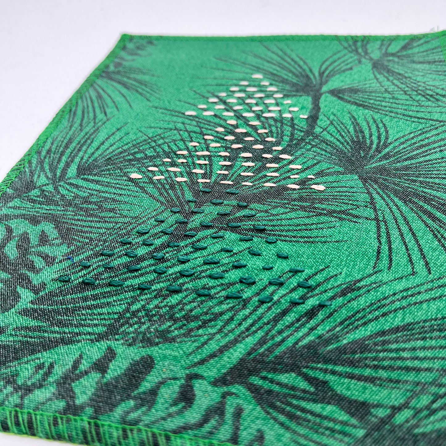 a fabric wall hanging made from bright green fabric with pine branches printed on it, and stitched over with Christmas trees made from three triangles, each triangle made from running stitches in green peach and ivory, on a white background