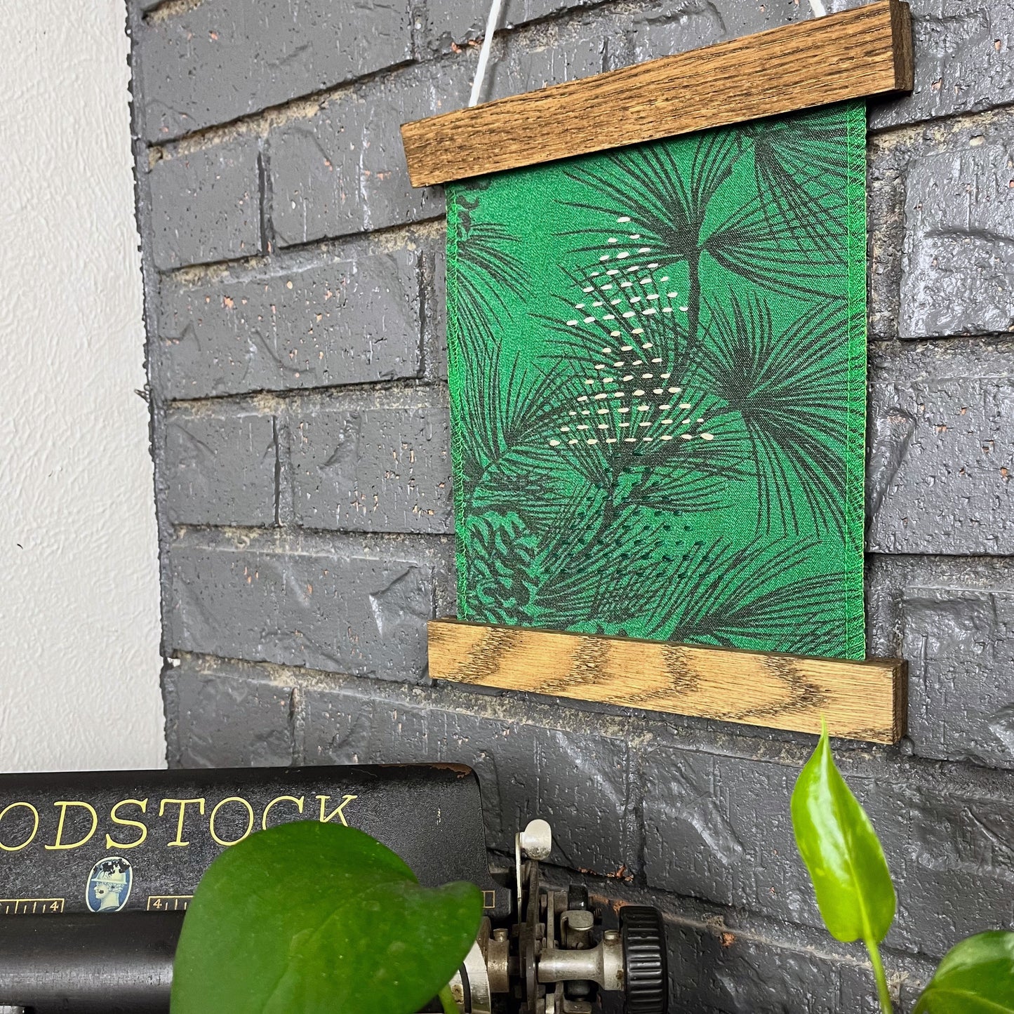 a fabric wall hanging in a magnetic wood frame, made from bright green fabric with pine branches printed on it, and stitched over with Christmas trees made from three triangles, each triangle made from running stitches in green peach and ivory