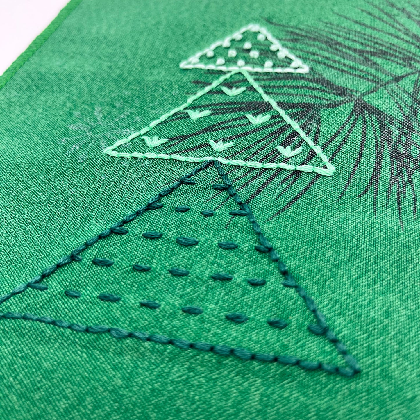 a close up view of a fabric wall hanging made from bright green fabric with pine branches printed on it, and stitched over with Christmas trees made from three triangles, each triangle filled with different types of stitches in shades of green