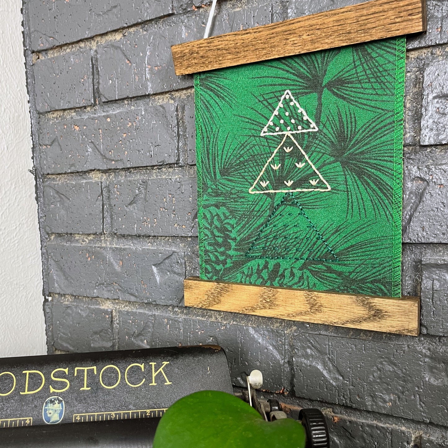 a fabric wall hanging in a magnetic wood frame, made from bright green fabric with pine branches printed on it, and stitched over with Christmas trees made from three triangles, each triangle filled with different stitches in green peach and ivory