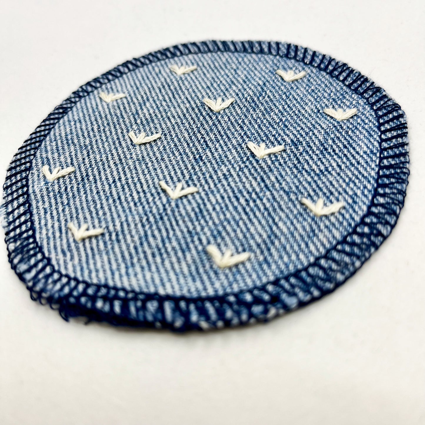 a close up angled view of a circle shaped patch made out of denim, with overlocked edges, with stitches in ivory that look like sprouts or birds feet, on a white background