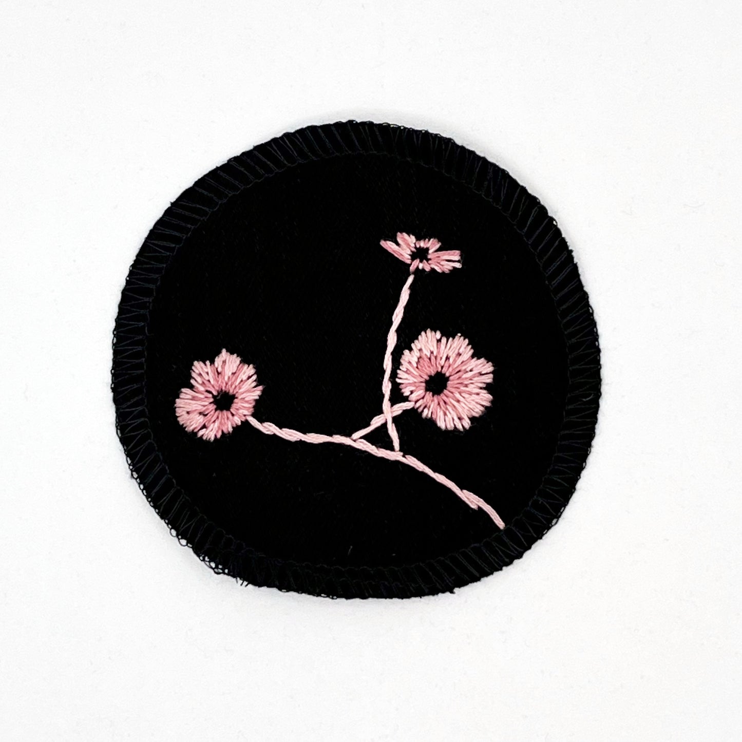 a circle shaped patch, made from black denim fabric, hand embroidered with 3 poppies in shades of pink, edges overlocked with black thread, on a white background