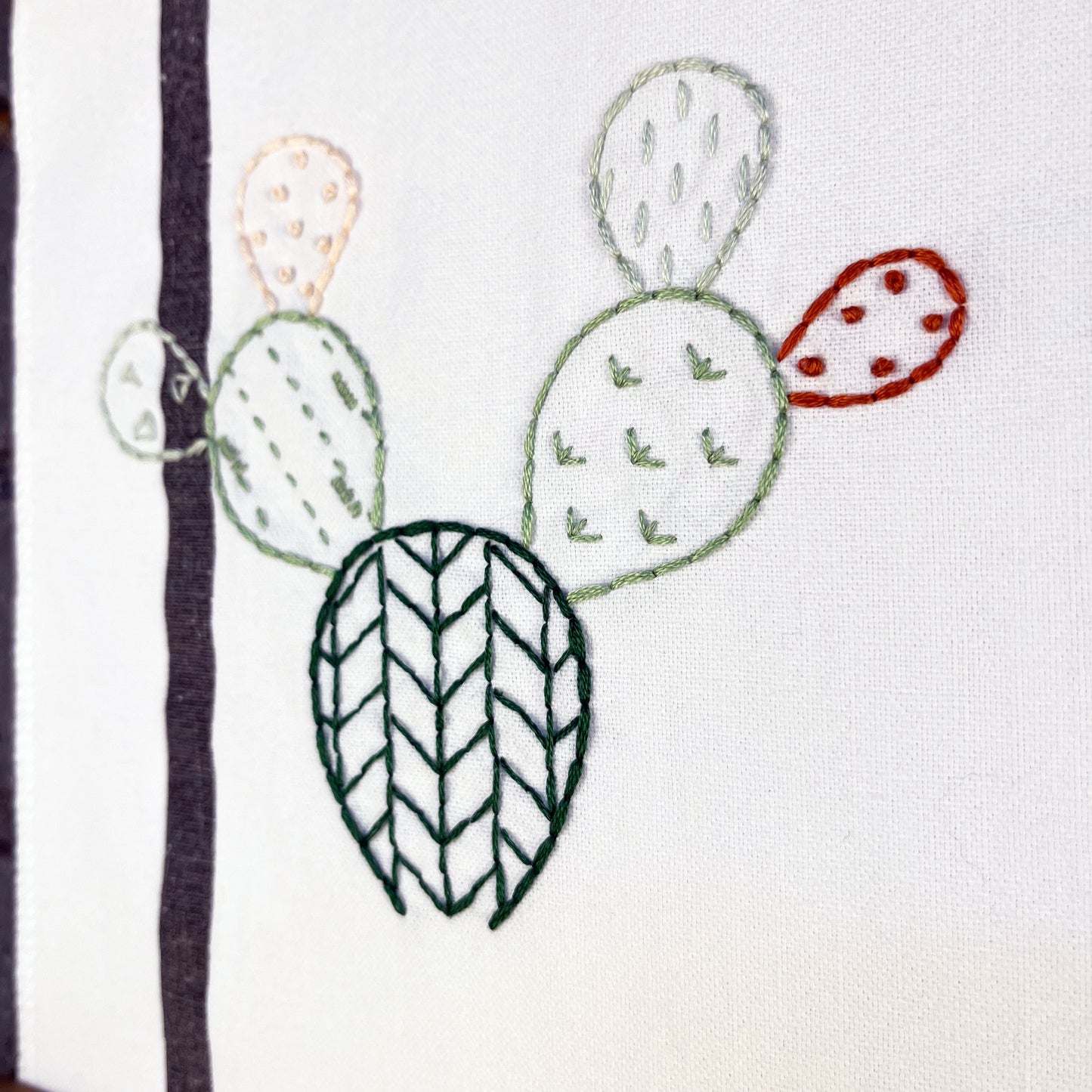 close up view of an outline of a prickly pear cactus hand embroidered in greens peach and rust brown on a white fabric with a black stripe, each pad filled with different types of stitches