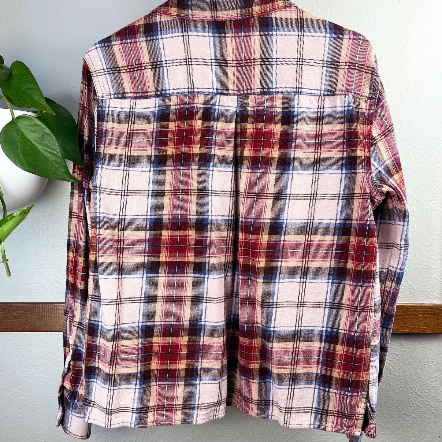 back view of a peach and maroon plaid flannel hanging on a hanger against a wall with a potted plant next to it