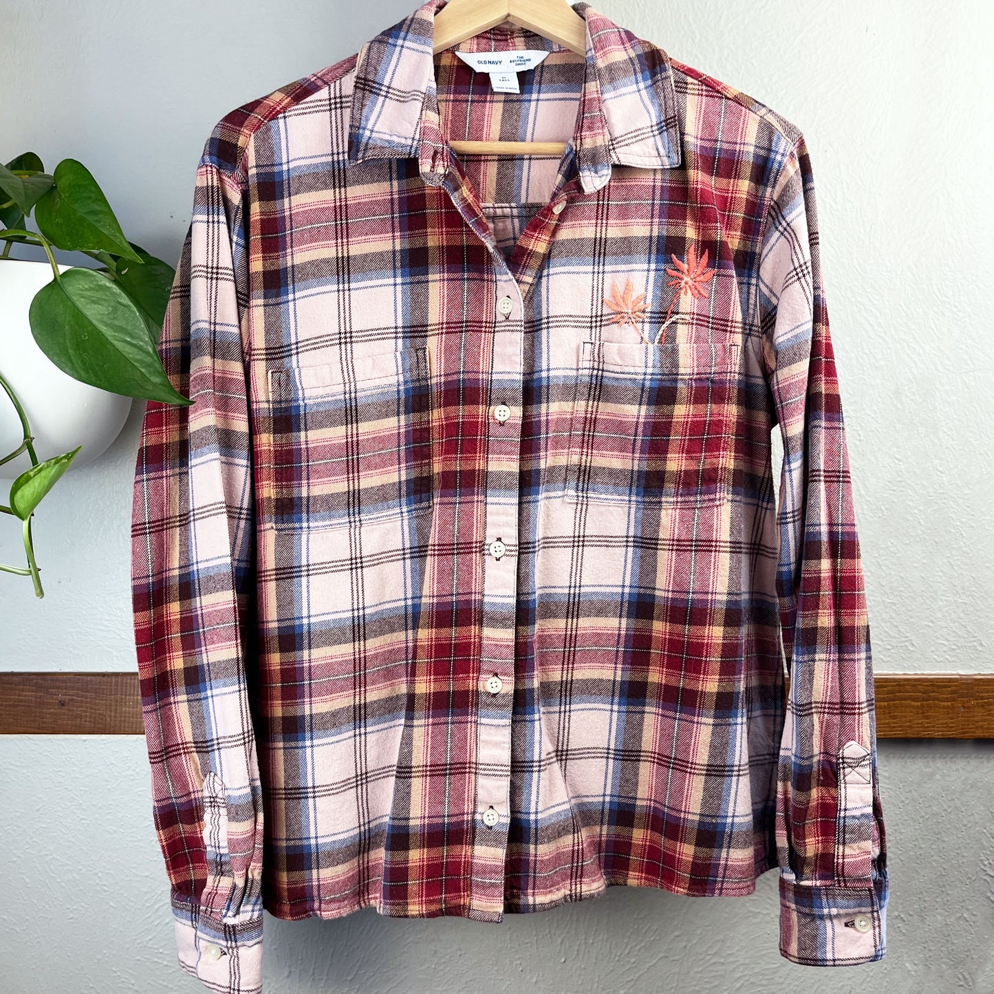 a peach and maroon plaid flannel with two hand embroidered flowers in peach and coral coming out of the chest pocket hanging on a hanger against a wall with a potted plant next to it