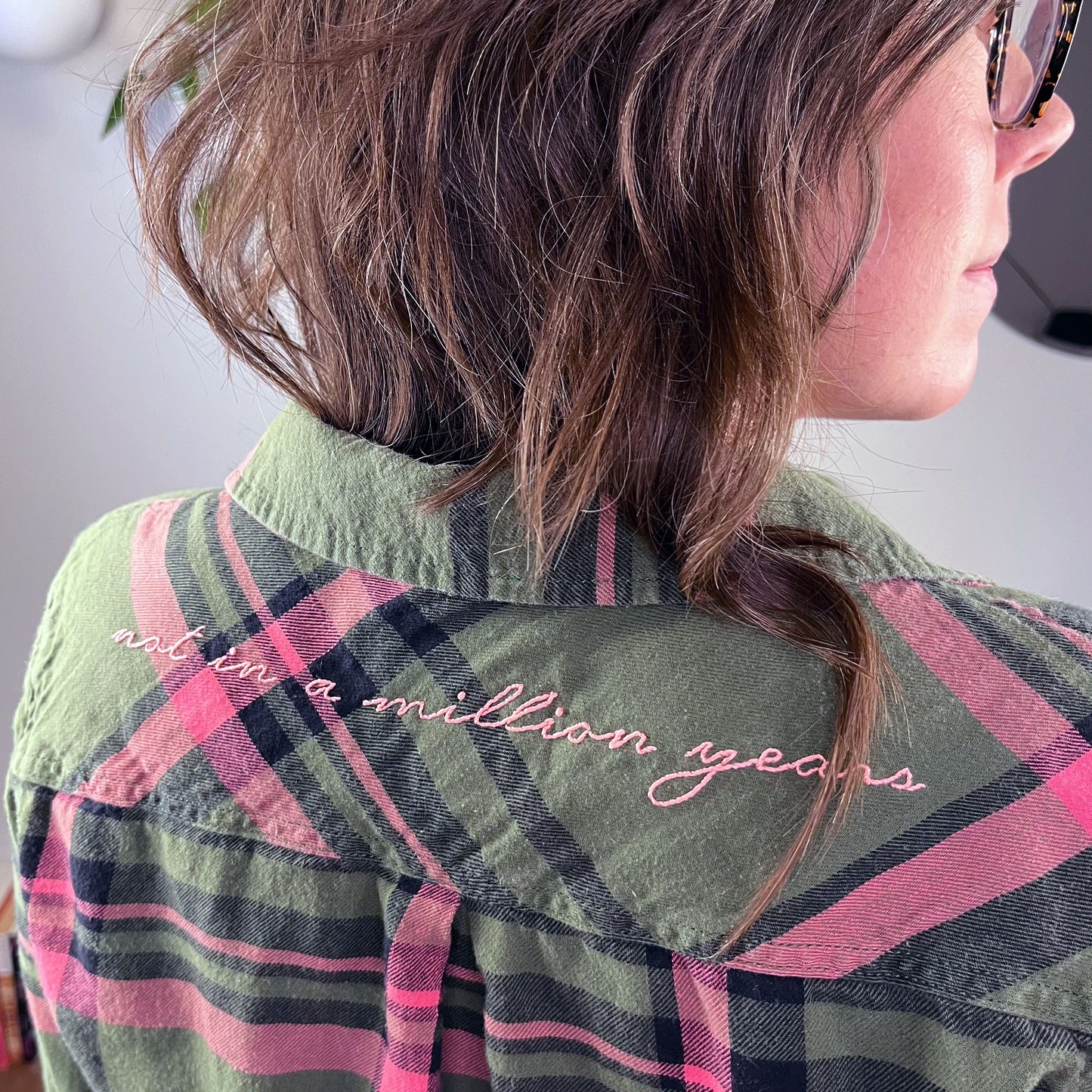 close up angled view of the back shoulders of a woman wearing a green and pink plaid flannel with the words not in a million years hand embroidered across the back yoke in pink scripty font