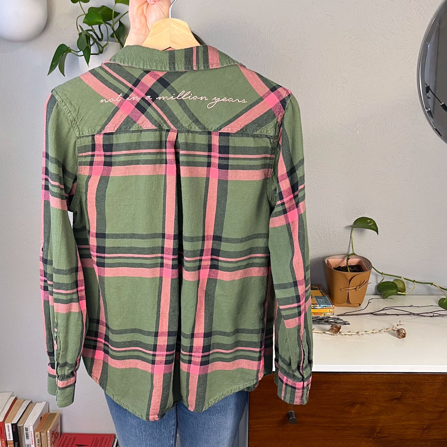 the back view of a green and pink plaid flannel hanging on a hanger with the words not in a million years hand embroidered across the back yoke in pink scripty font with books and plants in the background