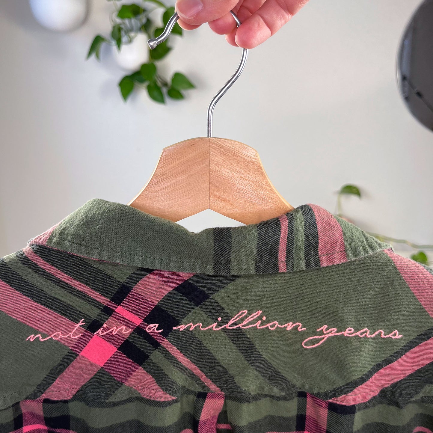 close up back view of a green and pink plaid flannel hanging on a hanger with the words not in a million years hand embroidered across the back yoke in pink scripty font with plants in the background