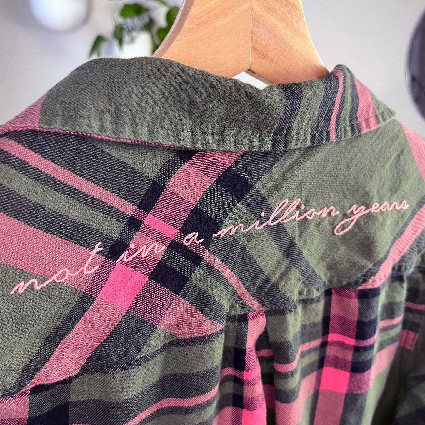 close up angled view of a back yoke of a green and pink plaid flannel with the words not in a million years hand embroidered across it in pink scripty font