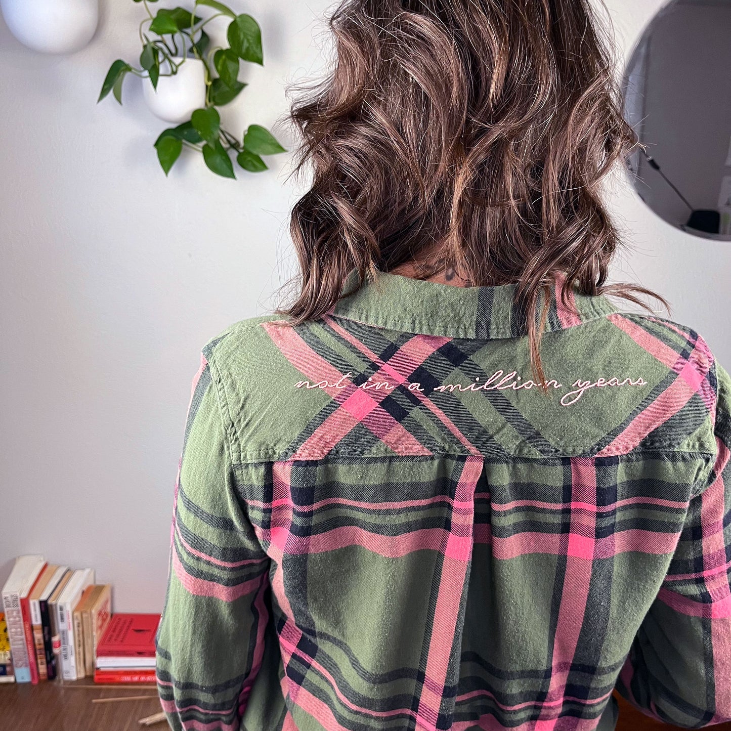 the back view of a woman wearing a green and pink plaid flannel with the words not in a million years hand embroidered across the back yoke in pink scripty font