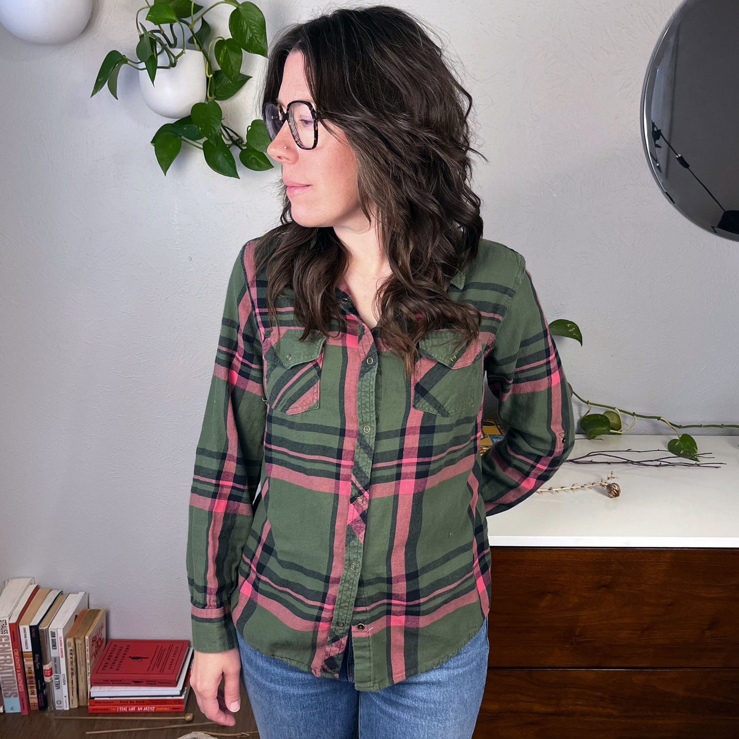 a white woman with glasses wearing a green and pink plaid flannel with books and plants in the background