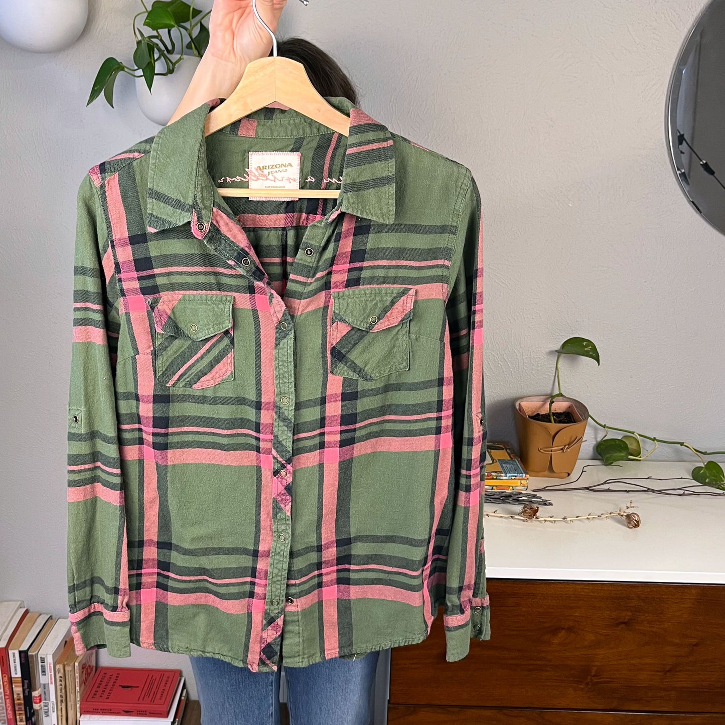 the front view of a green and pink plaid flannel hanging on a hanger with books and plants in the background