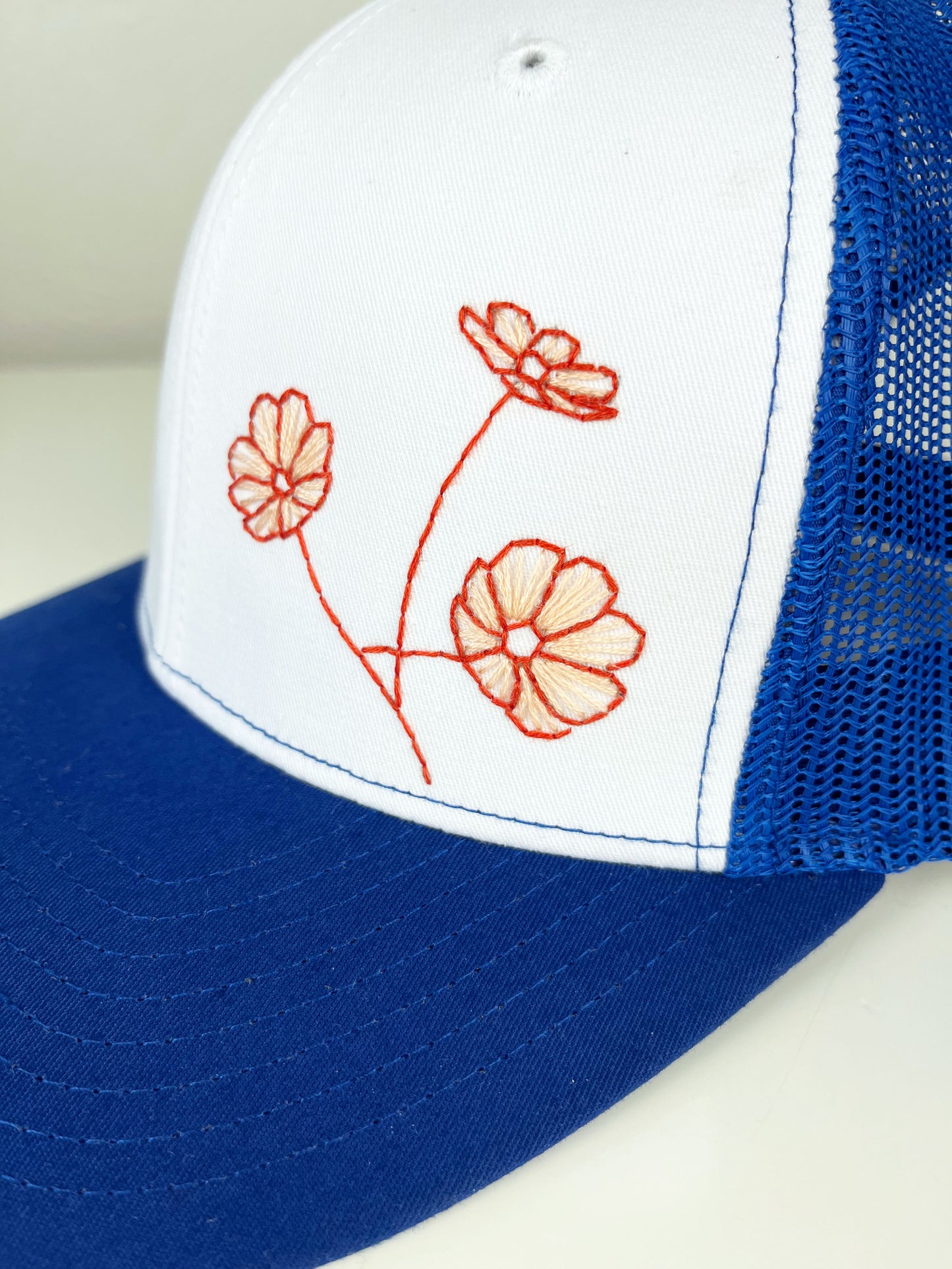 close up view of a blue mesh hat with poppies hand embroidered on the front in coral outline and peach line fill, sitting on a white counter top