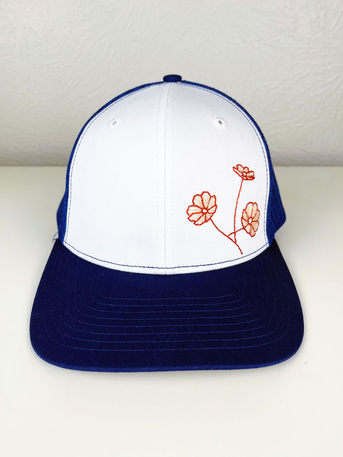 front view of a blue mesh hat with poppies hand embroidered on the front in coral outline and peach line fill, sitting on a white counter top