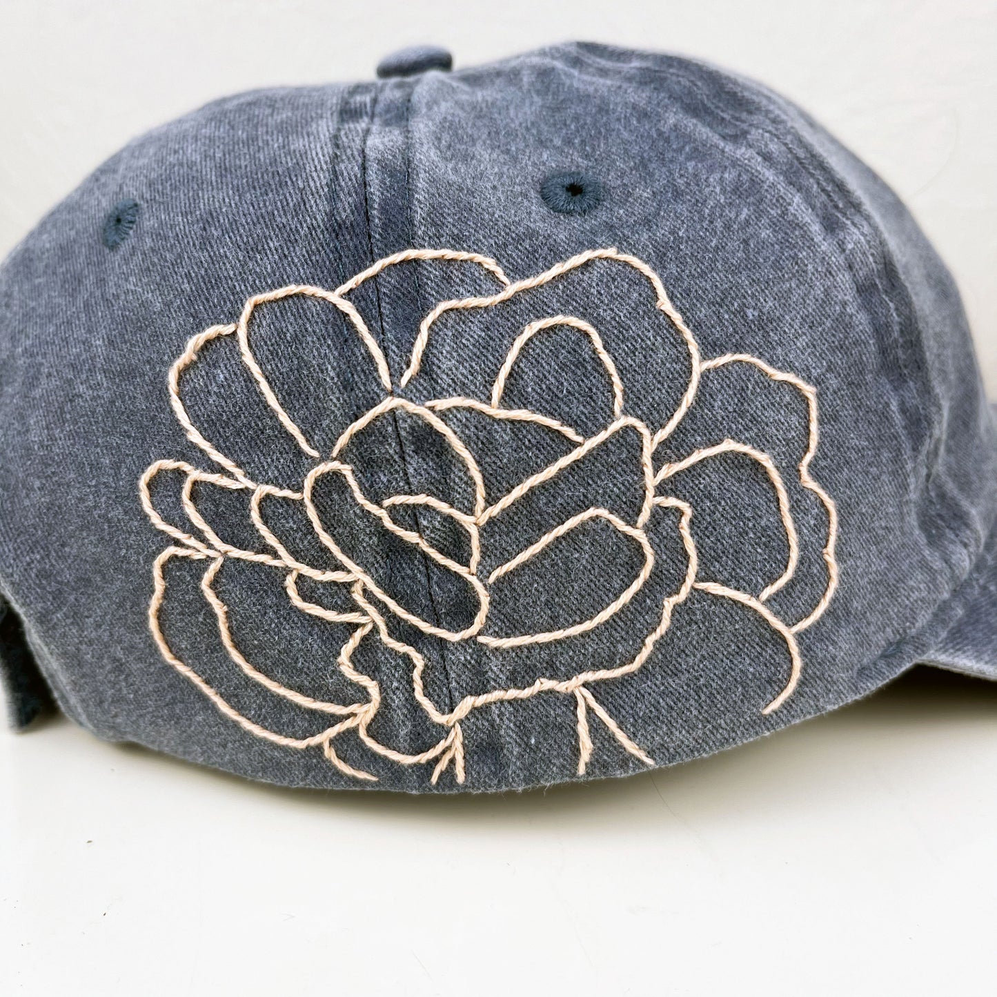detailed view of a large pale peach hand embroidered peony on the side of washed denim baseball hat sitting on a white dresser