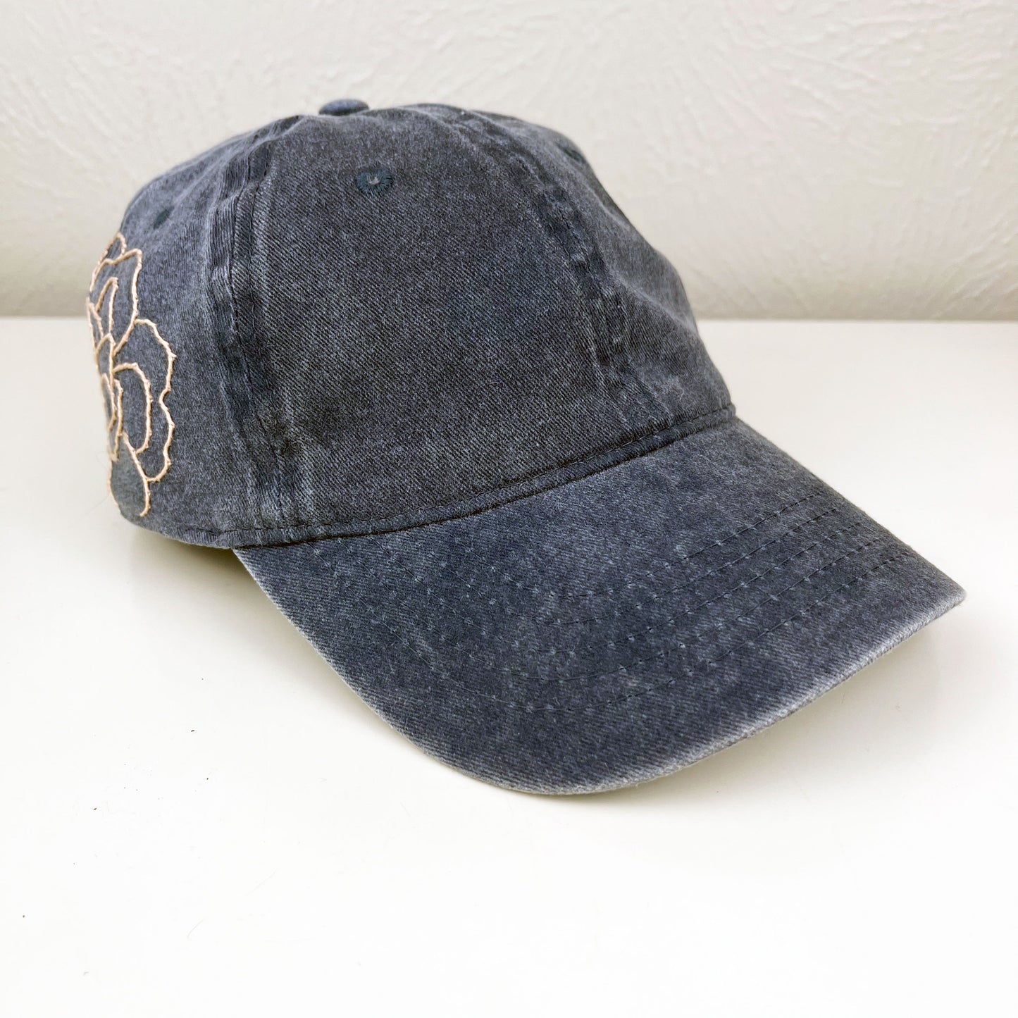 front angled view view of washed denim baseball hat with a large pale peach hand embroidered peony on the side, sitting on a white dresser