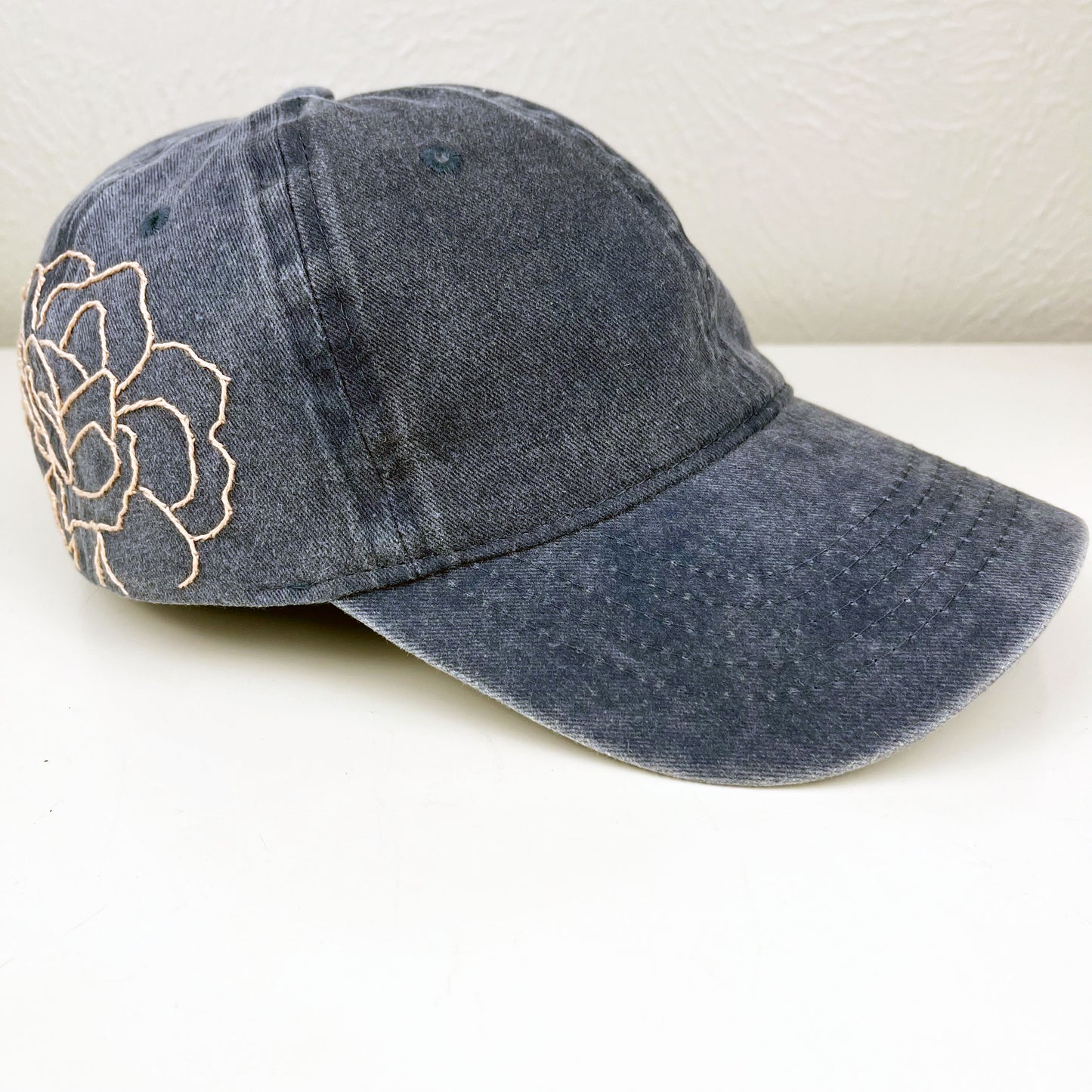 side view of washed denim baseball hat with a large pale peach hand embroidered peony on the side, sitting on a white dresser