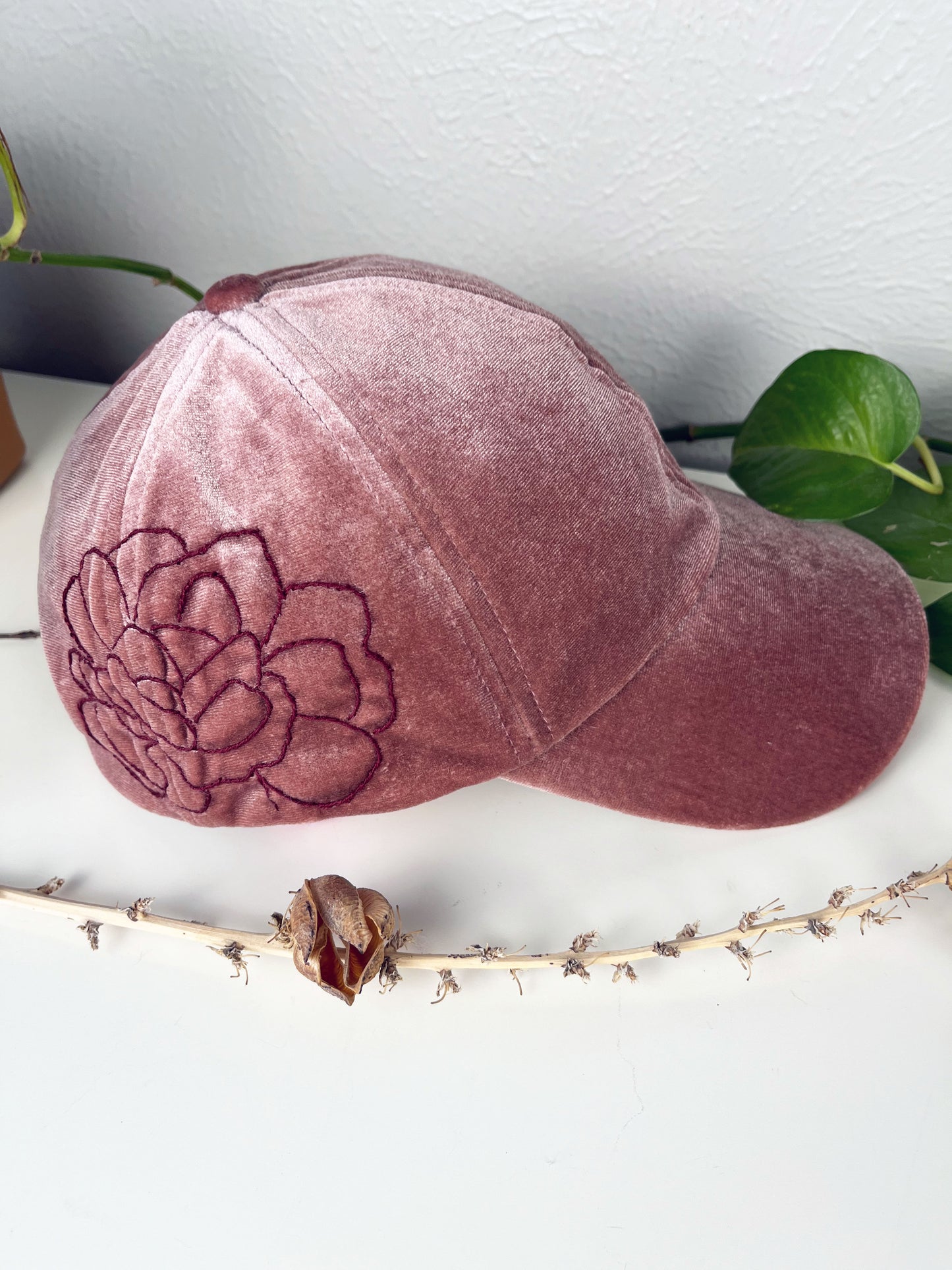 mauve velvet hat with dark mauve outline of a peony hand embroidered on the side, sitting on a white dresser with live and dried plants around it