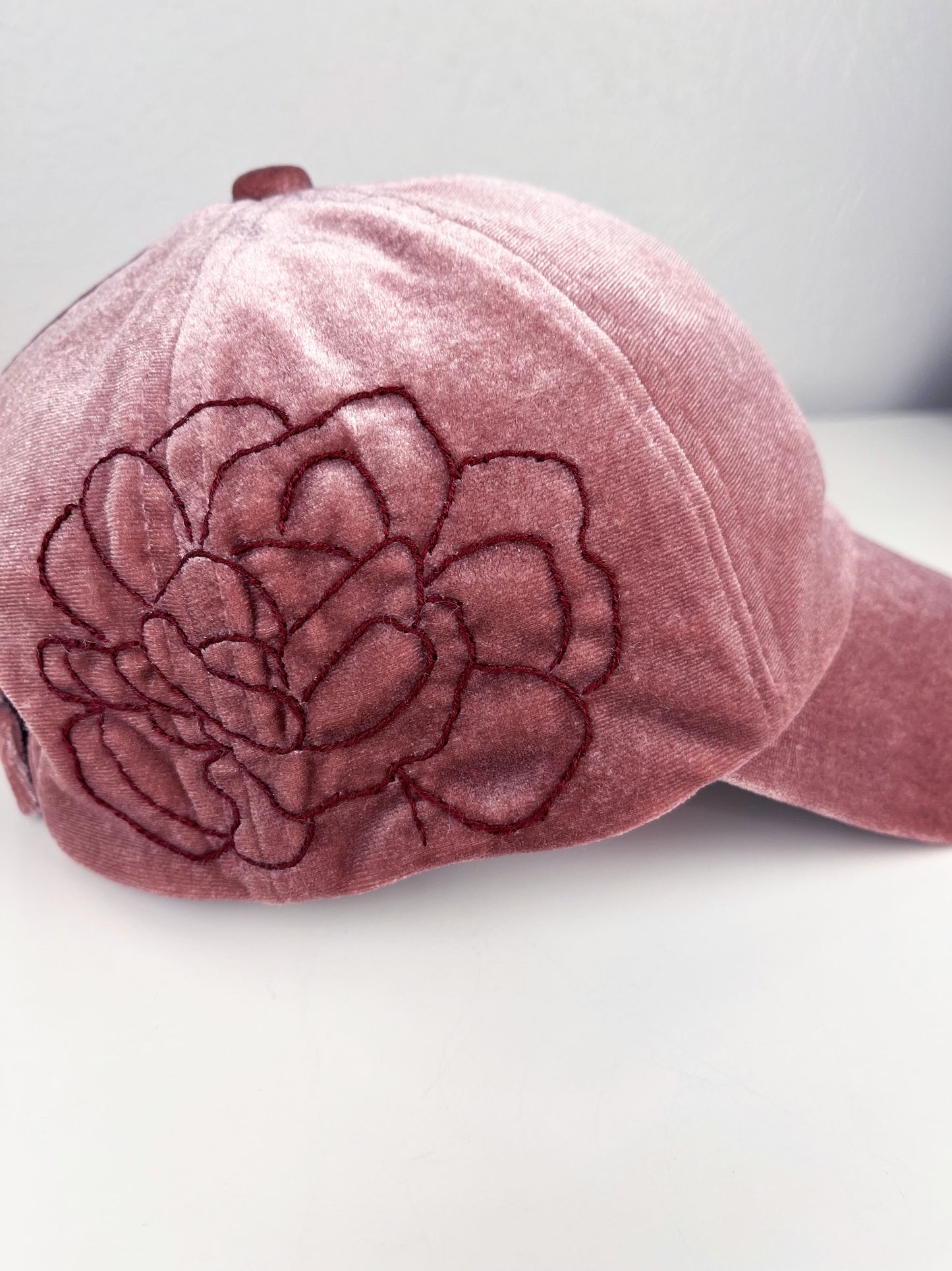 close up view of a mauve velvet hat with dark mauve outline of a peony hand embroidered on the side, sitting on a white dresser