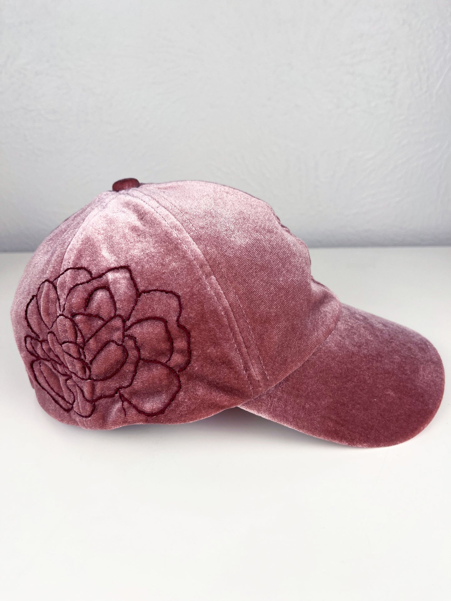 side view of a mauve velvet hat with dark mauve outline of a peony hand embroidered on the side, sitting on a white dresser