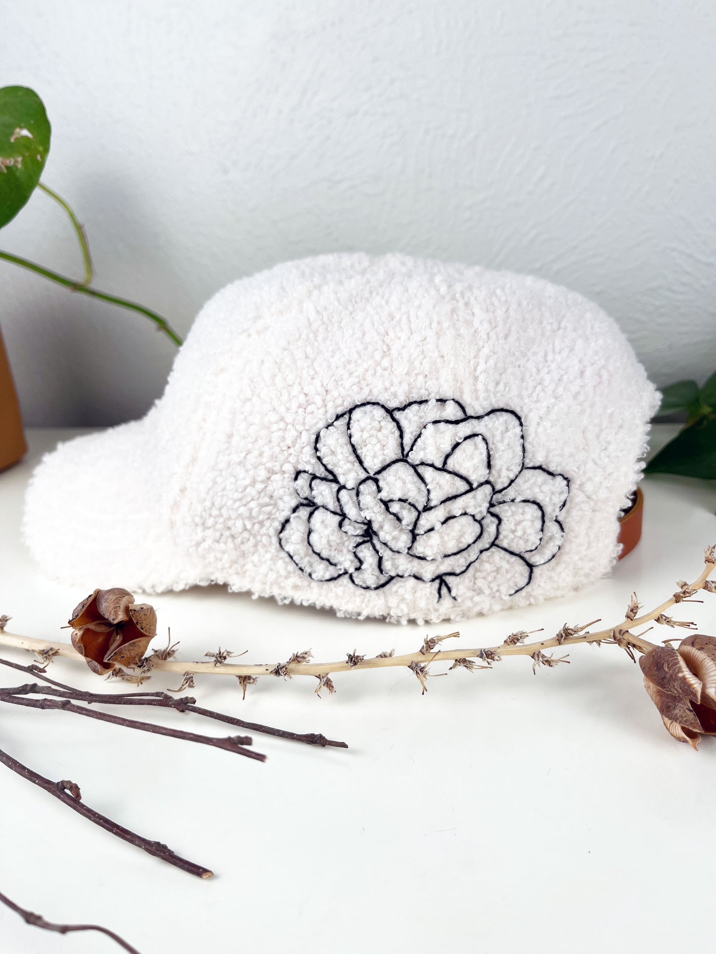 white boucle hat with black outline of a peony hand embroidered on the side, sitting on a white dresser with live and dried plants around it