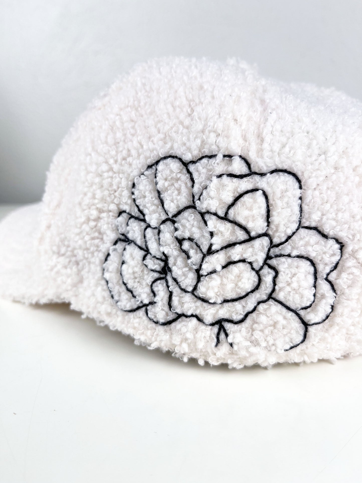 close up view of a white boucle hat with black outline of a peony hand embroidered on the side, sitting on a white dresser
