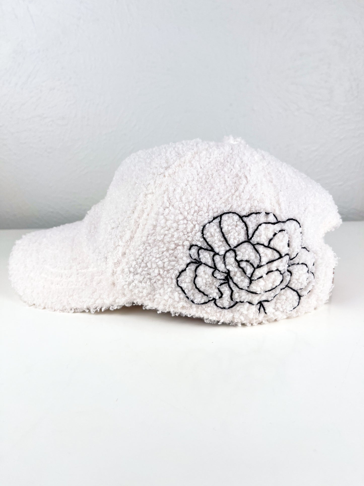 side view of a white boucle hat with black outline of a peony hand embroidered on the side, sitting on a white dresser