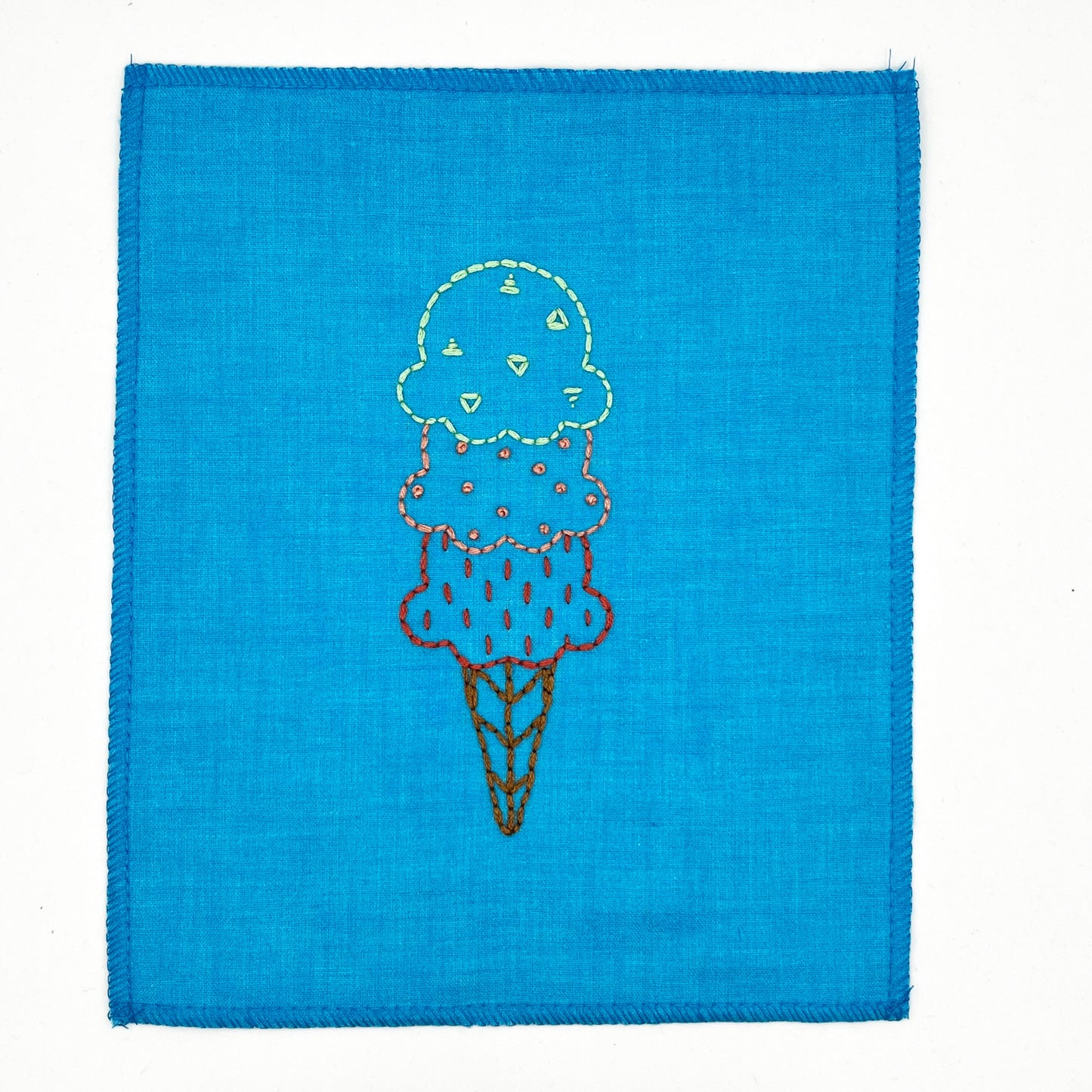 a small bright blue fabric wall hanging, hand embroidered with with scoops of ice cream on a cone, the top scoop outlined and filled with green triangles, the middle coral french dots, and the bottom red dashes, on a white background