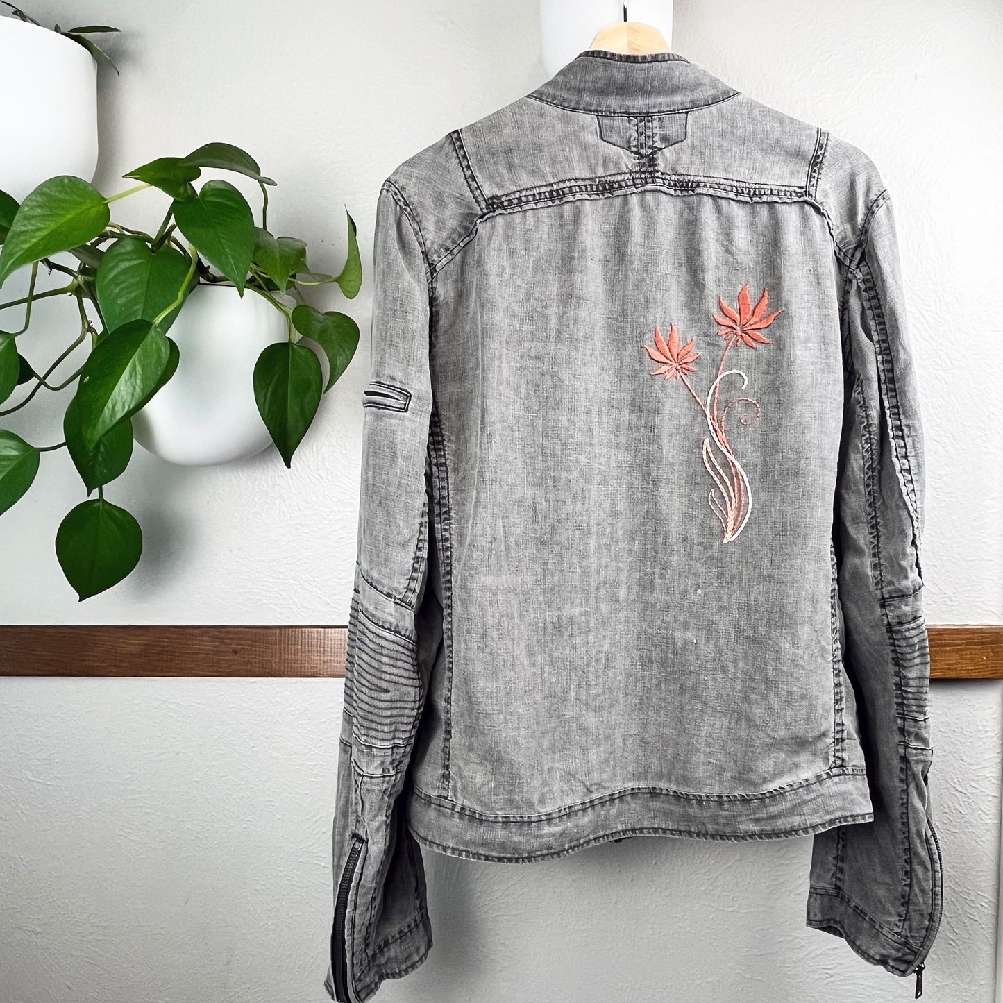 back of a grey linen moto jacket with long coral and peach flowers hand embroidered on the right side, hanging on a wall next to a pothos plant